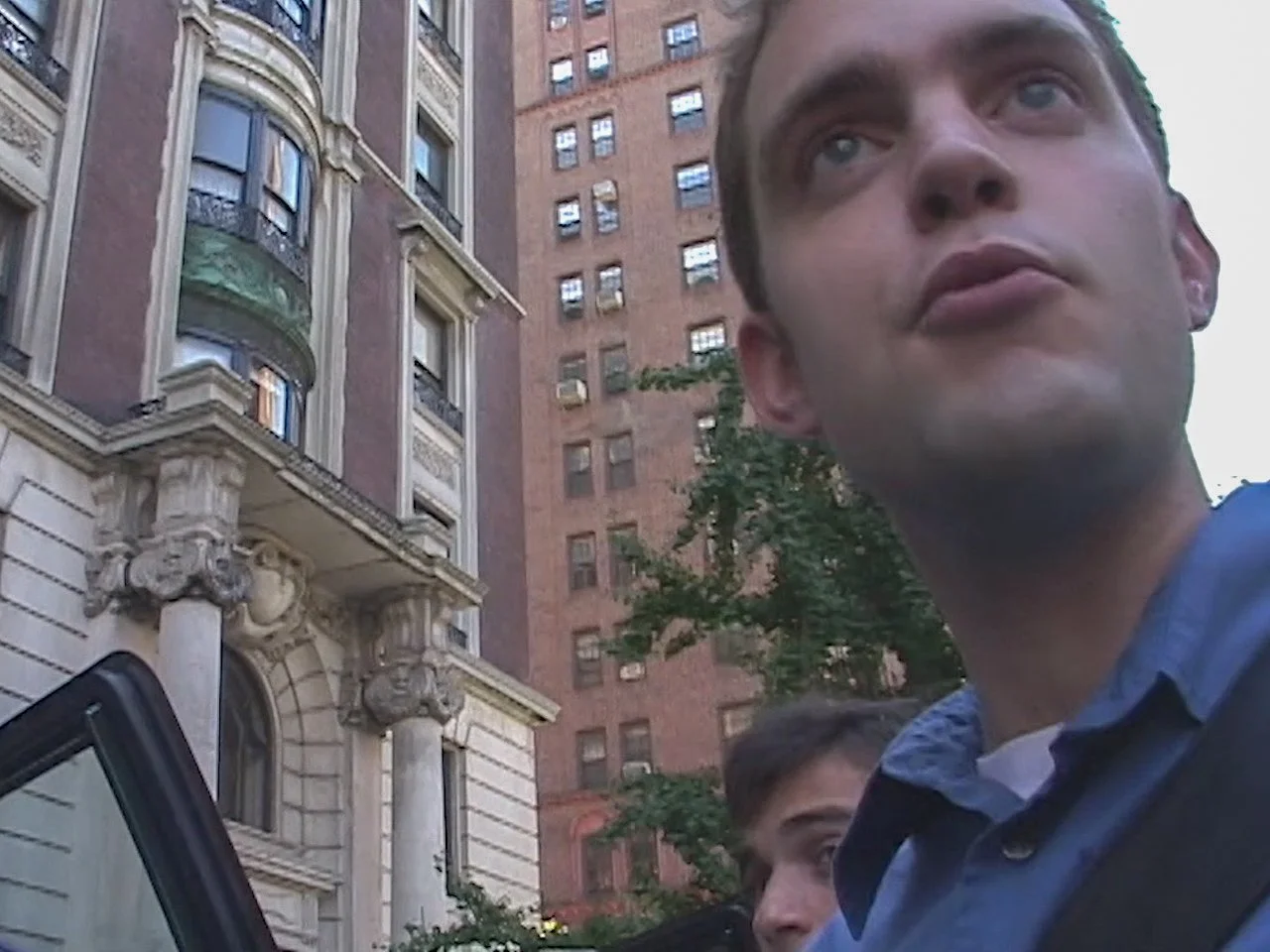 Close-up of a young man with short dark hair and blue eyes, wearing a collared shirt, standing outdoors in front of tall brick buildings with ornate architectural details. Part of another person with glasses is visible behind him.