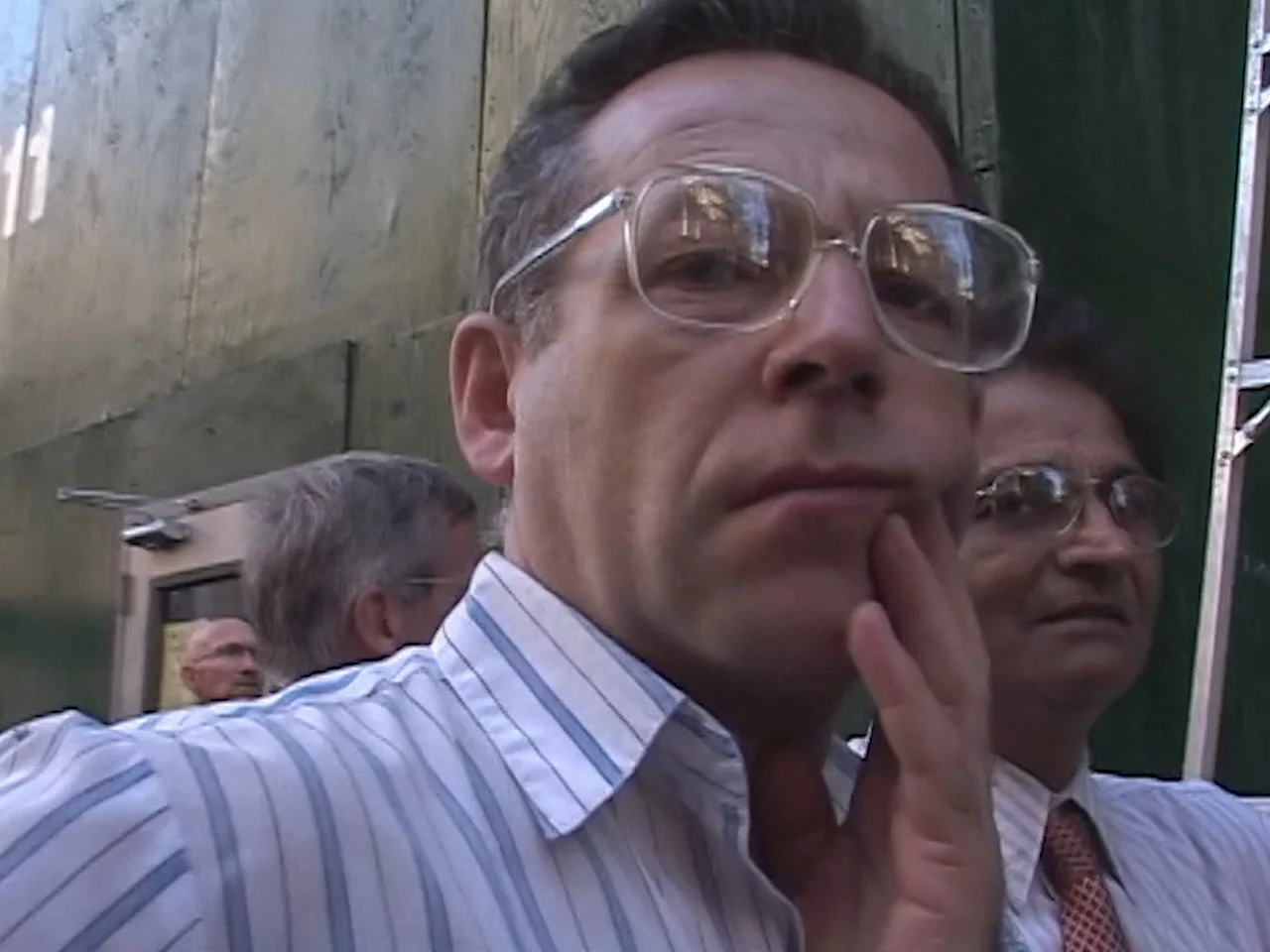 Close-up of a man with glasses touching his chin, with two other men in glasses and a man in the background, outdoors near a wooden structure and scaffolding.   New York City.   9/11 September 11th 2001