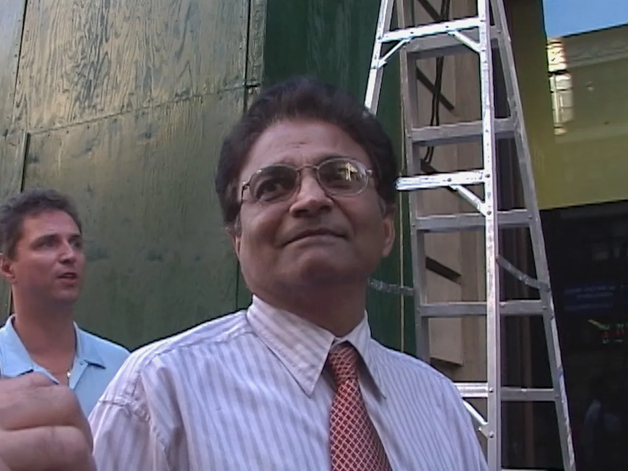 A man with glasses wearing a striped shirt and red tie smiling outdoors, with another man in the background and a metal ladder beside them.   New York City.   9/11 September 11th 2001