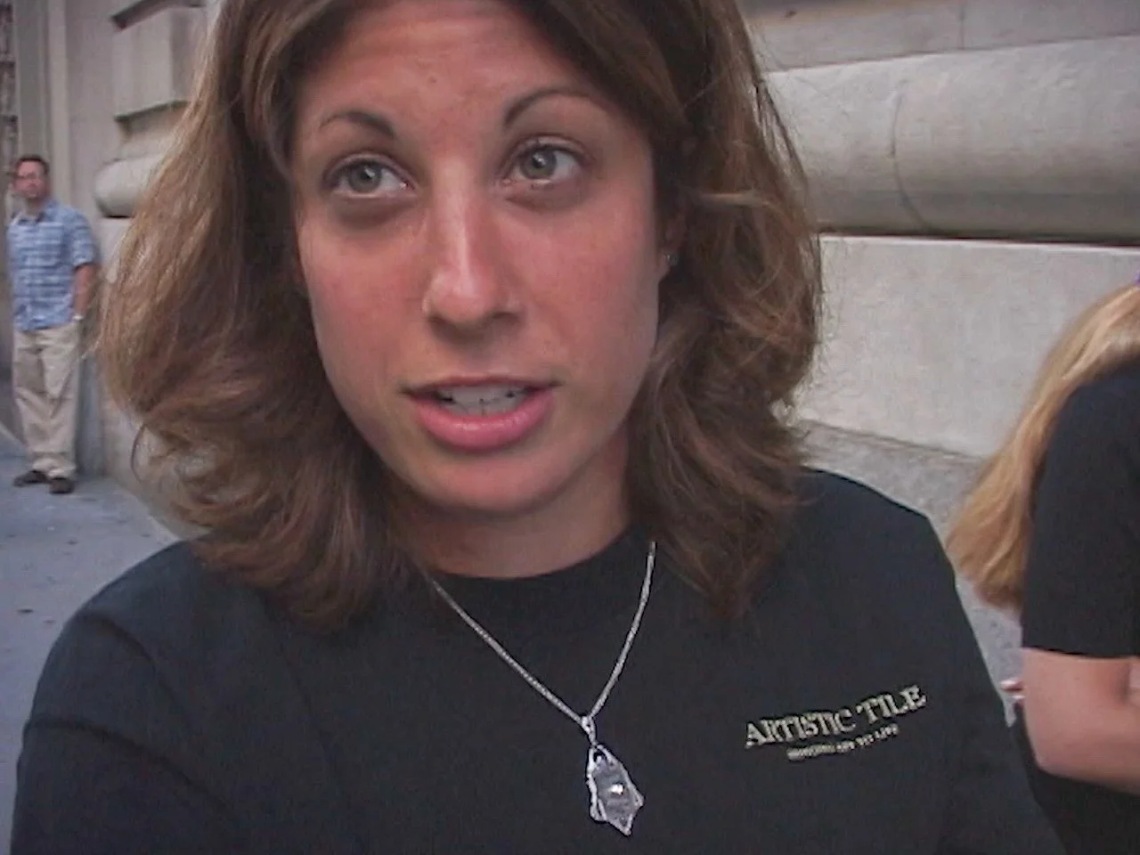 A woman with shoulder-length brown hair and fair skin, wearing a black shirt with a logo, and is talking outdoors in front of a stone wall. She is wearing a necklace with a clear pendant.