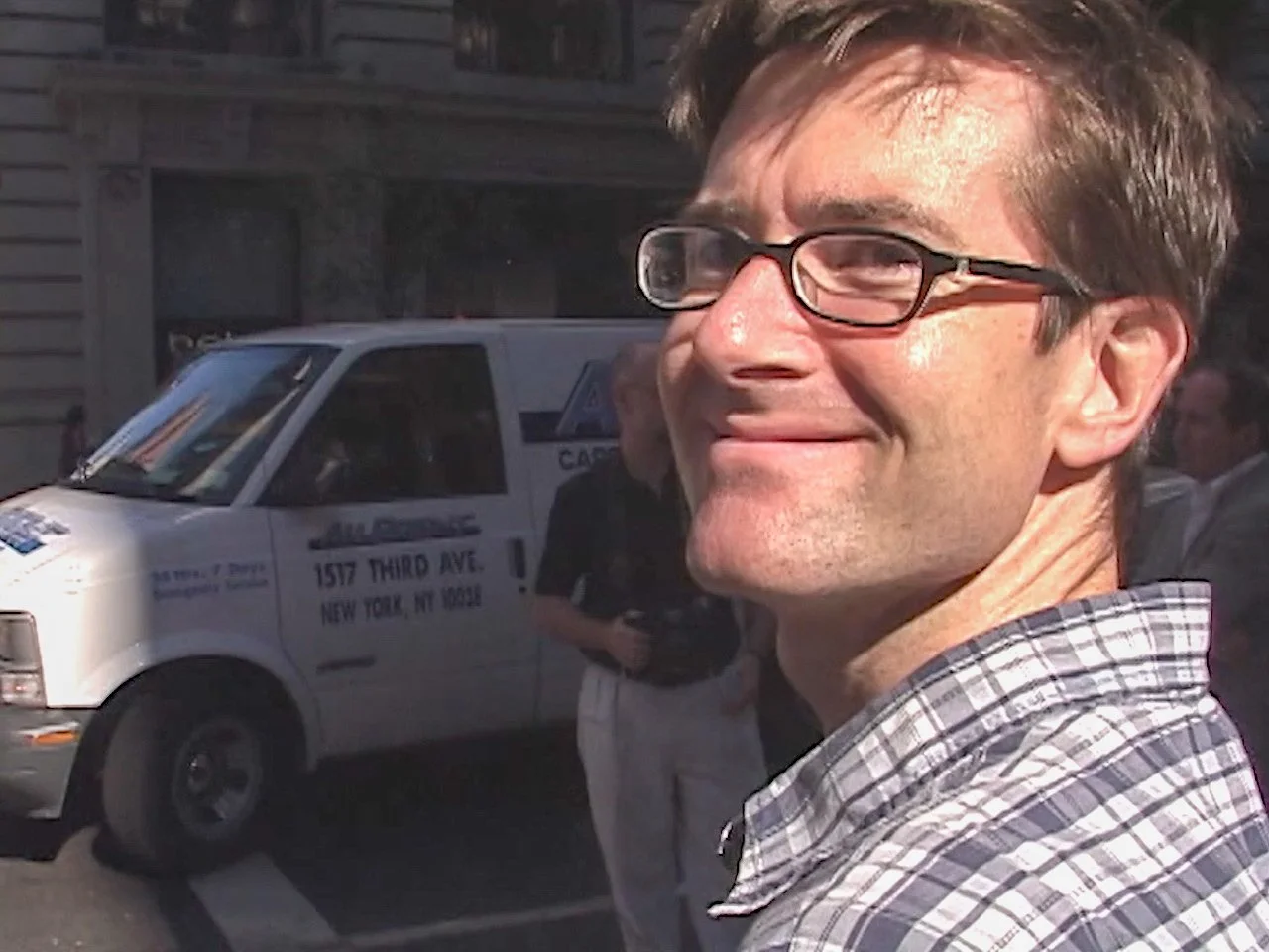 A man wearing glasses smiling in an outdoor city street, with a white van and other people in the background.