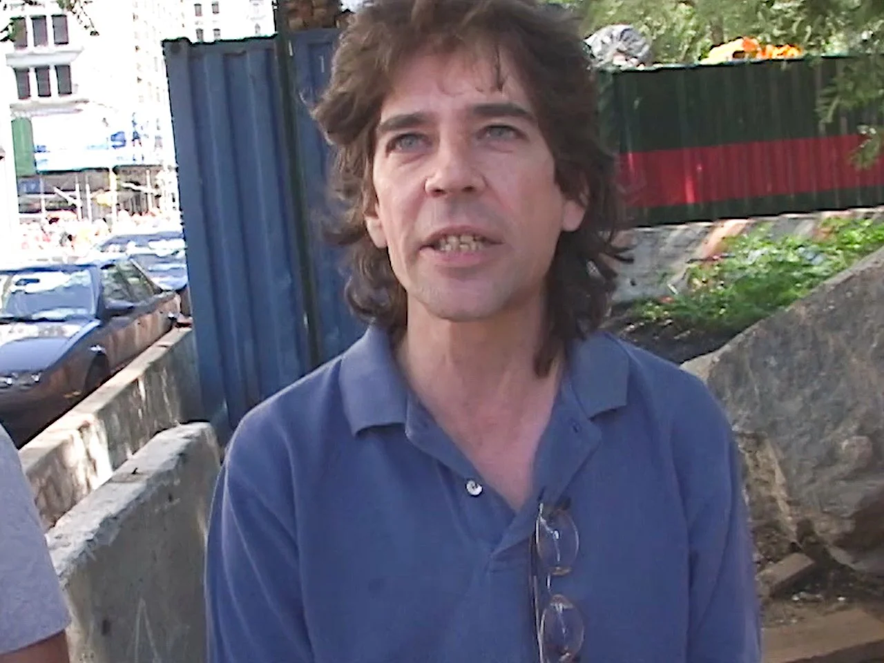 A man with curly brown hair and blue eyes wearing a blue polo shirt, standing outdoors in an urban environment with parked cars and construction fencing in the background.