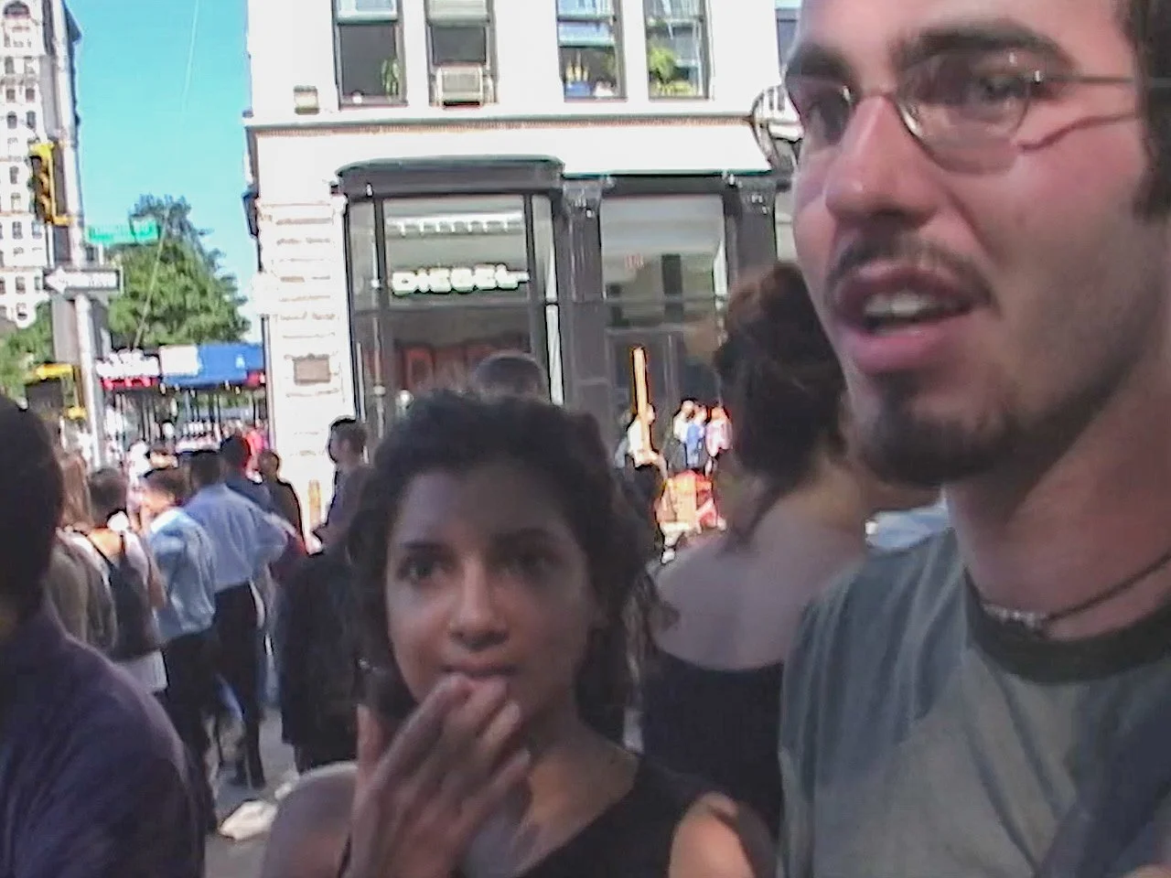A crowded city sidewalk with buildings and a bus in the background. People are walking and socializing, including a woman touching her lips and a man wearing glasses and a t-shirt, both in the foreground.
