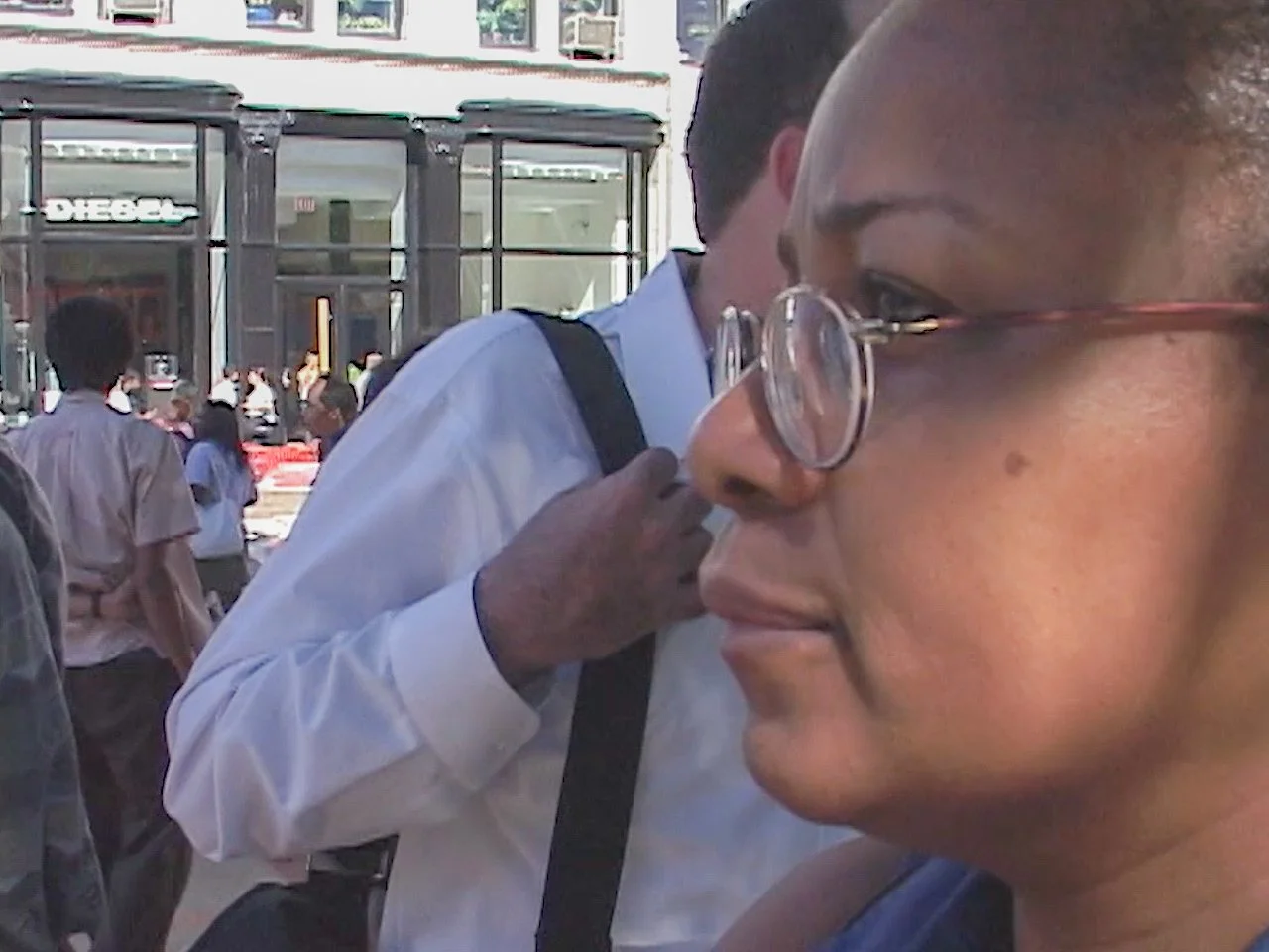 Close-up of a woman wearing glasses in a busy outdoor urban setting with people and storefronts in the background.
