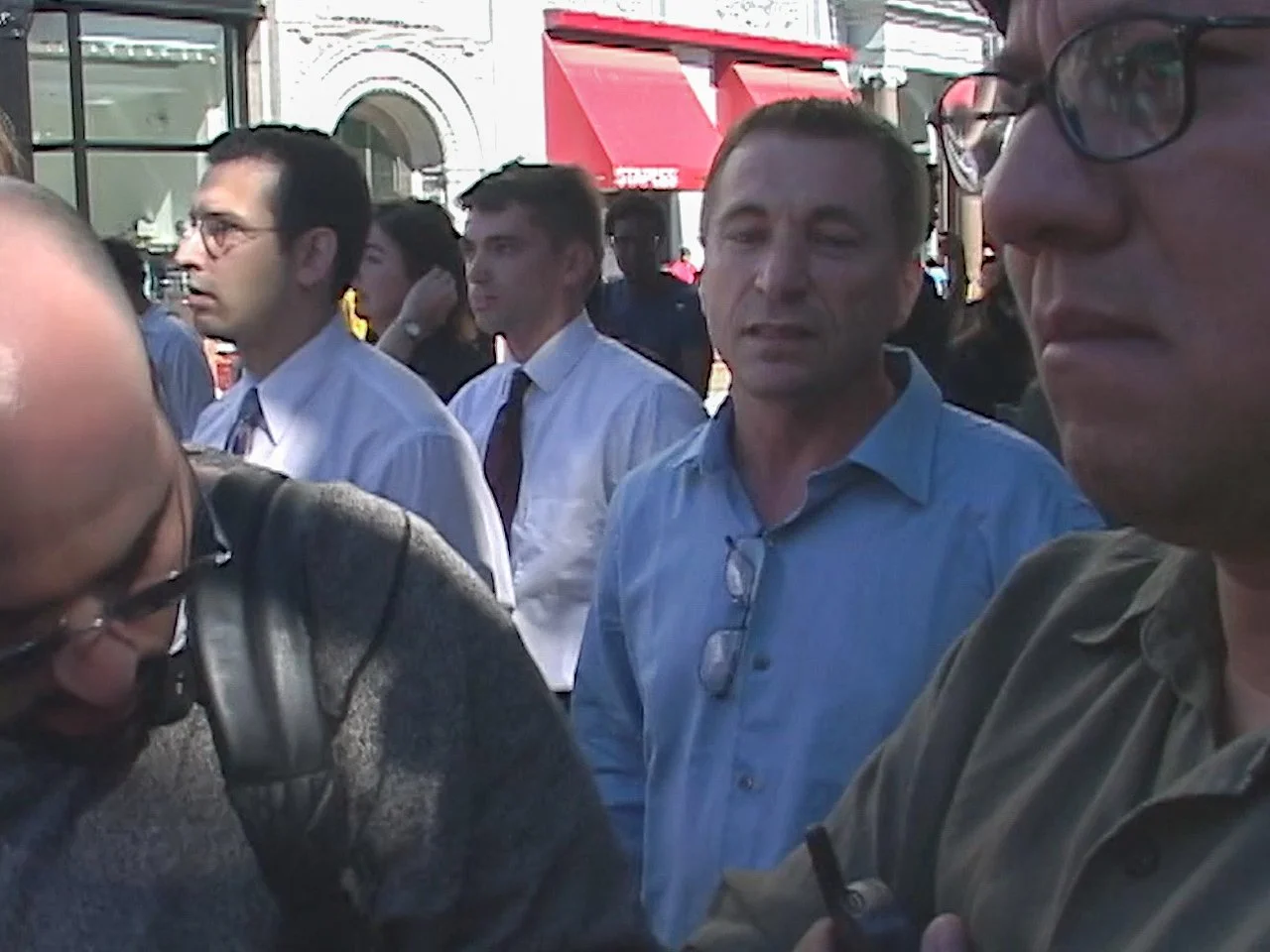 Group of people gathered outdoors, some wearing glasses and formal shirts, engaging in conversation, with city buildings and a red canopy in the background.