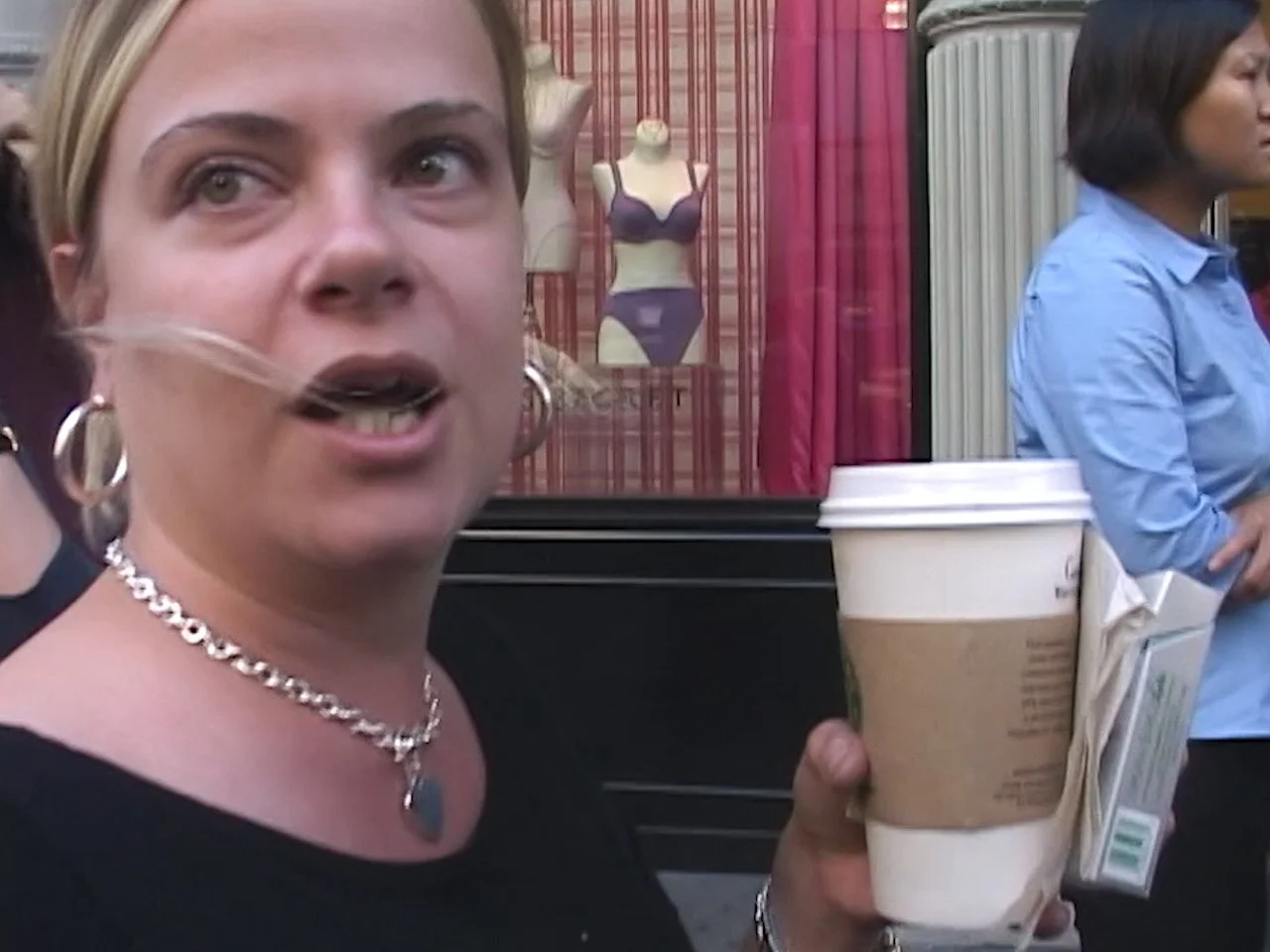 A woman with blonde hair and hoop earrings talking, holding a large cup of coffee with a cardboard sleeve, outside on a street with a shop window and mannequins in the background. 9/11 September 11th 2001