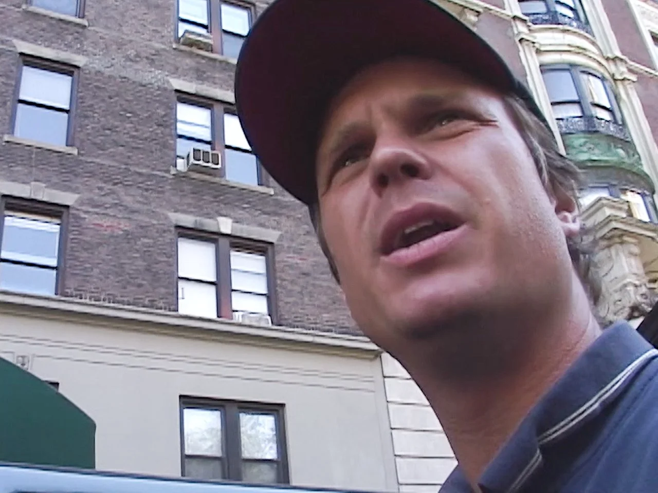 Close-up of a man wearing a baseball cap, speaking or singing with an urban building in the background.