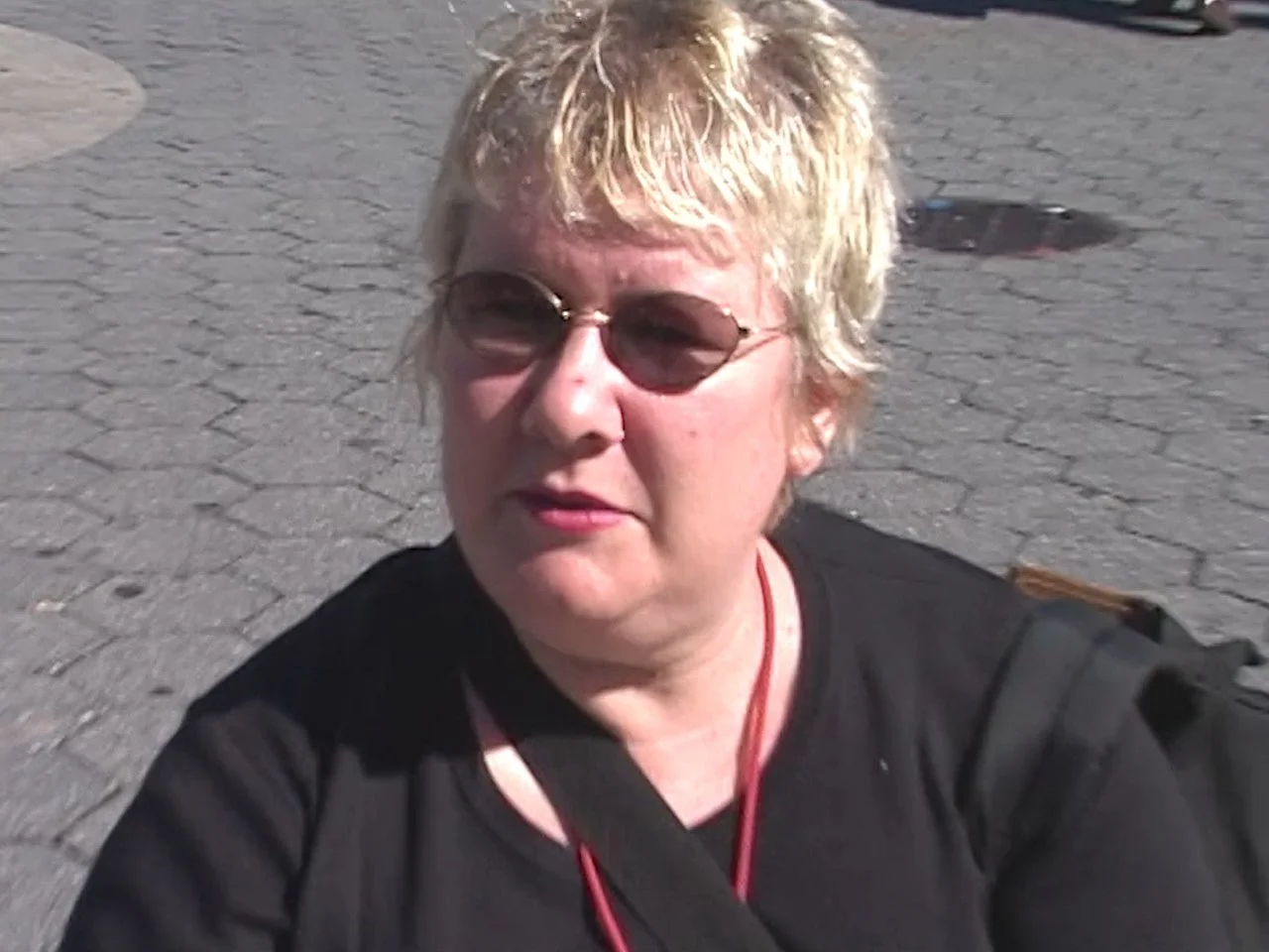 A woman with short, blonde hair wearing sunglasses and a black shirt, sitting on a paved surface outdoors.