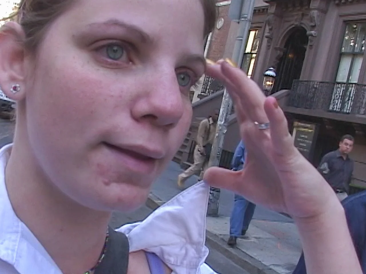 Close-up of a woman with light skin, freckles, and blue eyes, touching her head with her hand, standing on a city street with buildings and other pedestrians in the background.