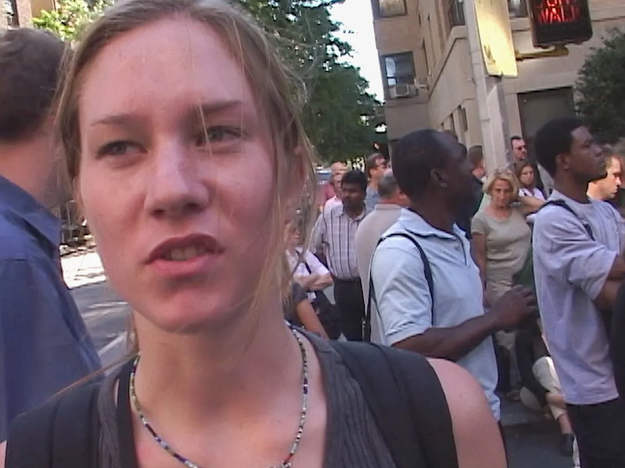 A woman with light brown hair and a beaded necklace standing among a crowd on a city street during daylight.