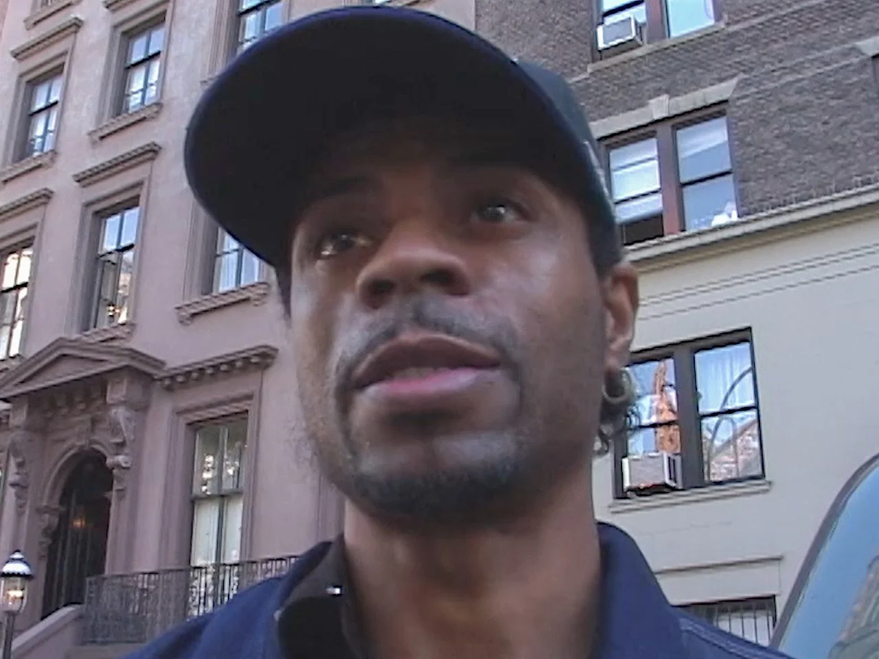 Close-up of a man wearing a cap, standing outdoors in front of a building with multiple windows.