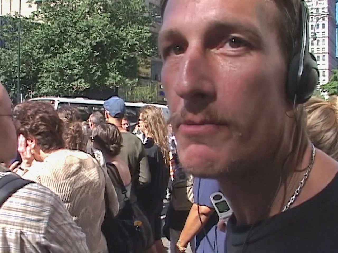 Close-up of a man wearing headphones in a crowded outdoor setting, with trees and buildings in the background.