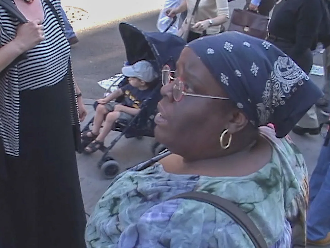 A close-up of a woman wearing a bandana, glasses, and hoop earrings, standing outdoors in a crowd. A child in a stroller is visible in the background, along with several other people.