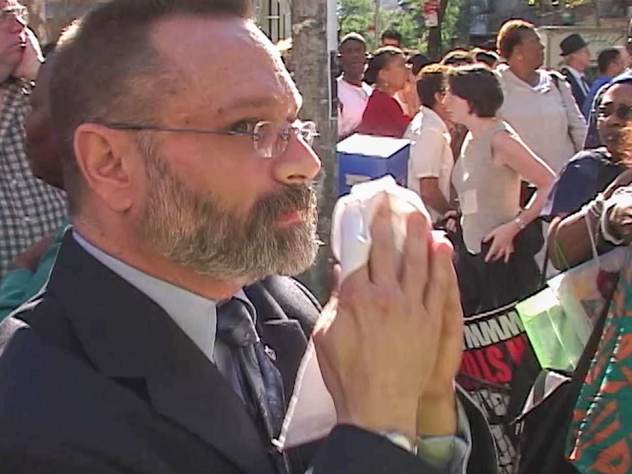 A man with glasses and a beard is praying with his hands pressed together, surrounded by a crowd of people outdoors.