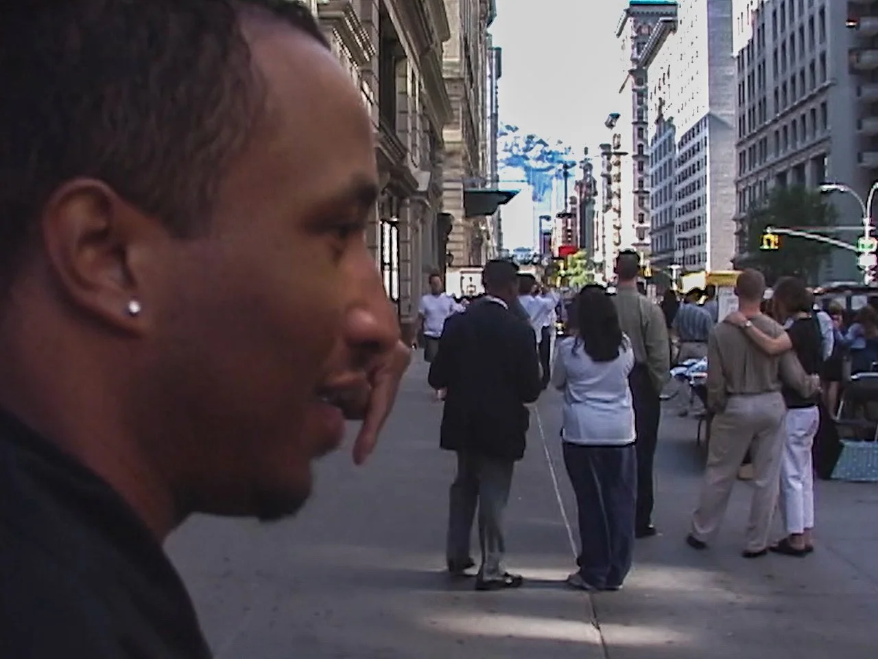 A man with short hair, earrings, and a goatee is in the foreground of a busy city street, with people walking and talking among tall buildings, yellow taxis, and streetlights.    New York City.   9/11 September 11th 2001