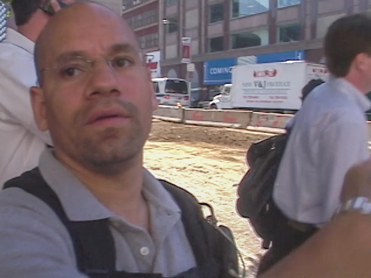 A man with glasses and a backpack taking a selfie outdoors in an urban area with construction site in the background.