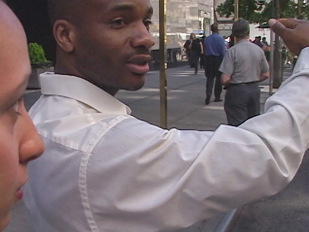 A man in a white shirt gesturing or talking on a busy city street, with other pedestrians walking in the background.