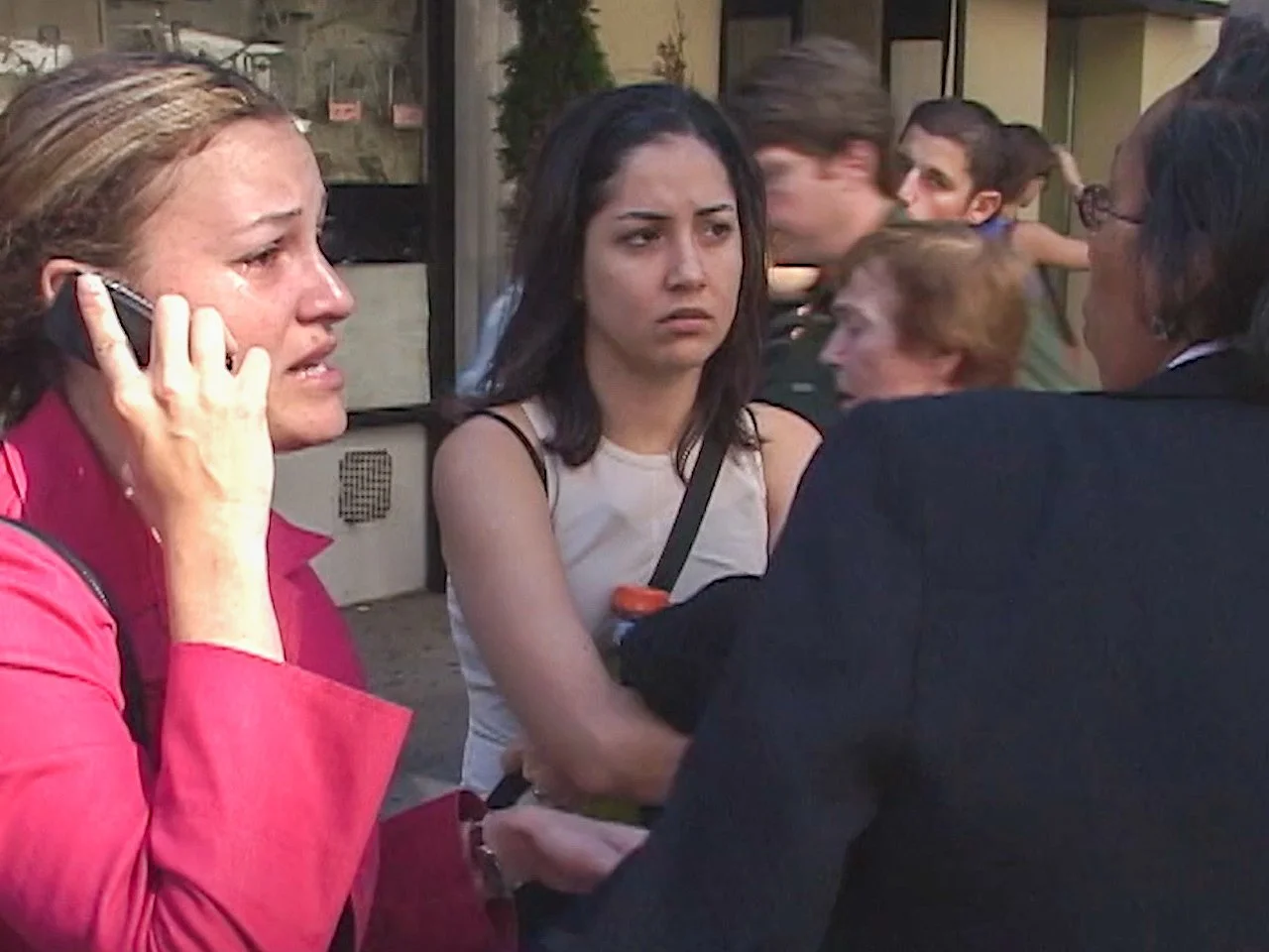 Three women and a man are engaged in a conversation outside. One woman with blonde hair on the left is on the phone, another woman with dark hair in the middle appears worried, and the man on the right has glasses and dark hair. Other people are blur