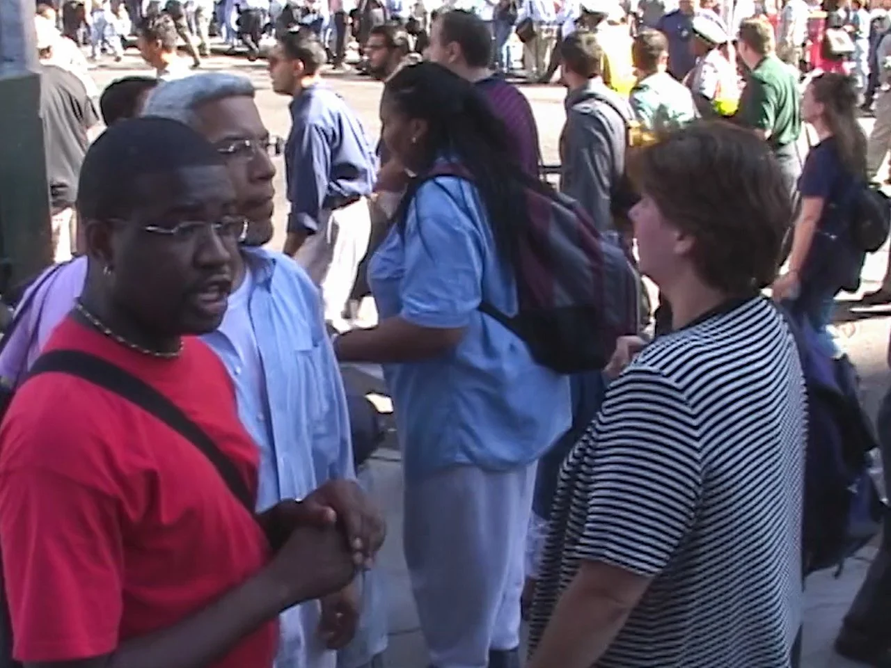 People congregate outdoors in a crowd, engaging in conversation. The scene depicts a diverse group of individuals standing and talking on a sunny day.