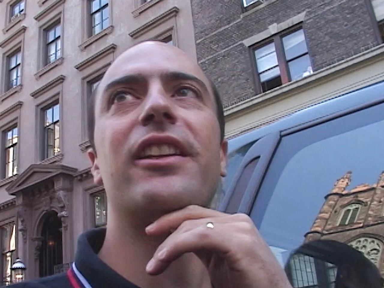 Close-up of a smiling man with short hair, wearing a dark jacket with red and white accents, standing outdoors in a city street with brick and stone buildings and a parked vehicle in the background.