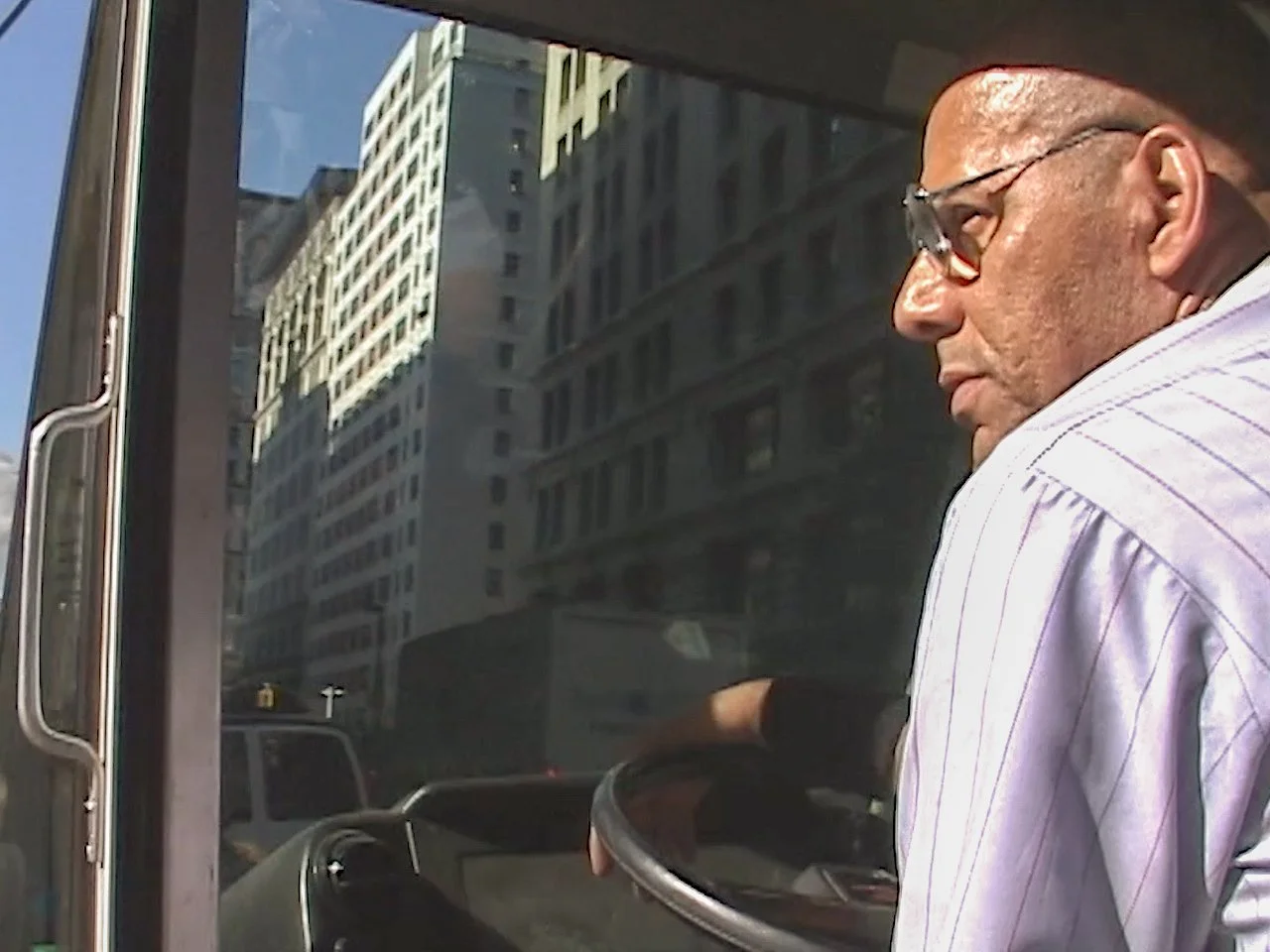 A man with glasses wearing a white striped shirt standing next to a glass window or door in an urban city setting with tall buildings reflected in the glass.