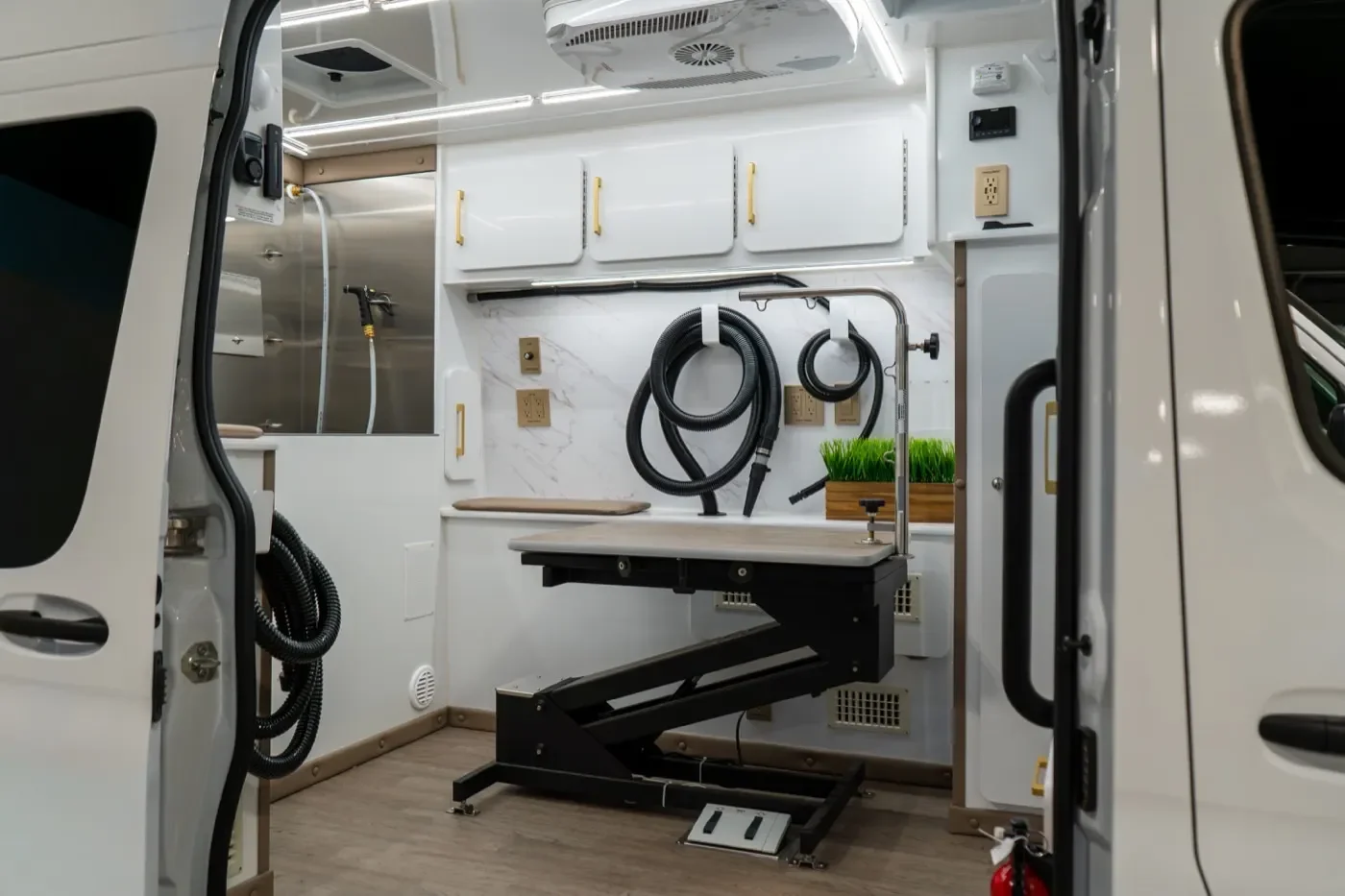 Interior of a high end mobile grooming van with adjustable grooming table, organized with storage cabinets, pet dryer hoses, a bath tub, and a small potted plant.