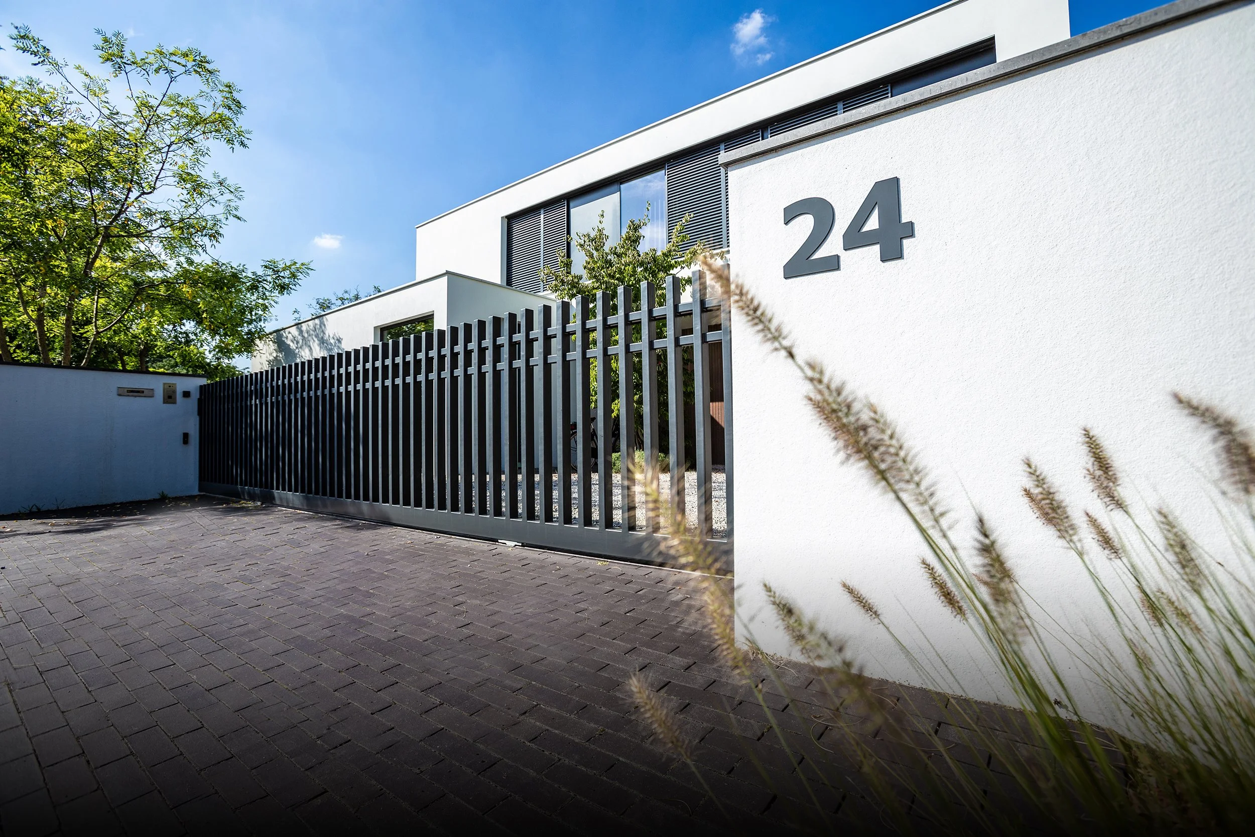 Wit modern huis met zwart getal 24 op de muur, omgeven door een zwarte metalen poort, groene bomen en een heldere blauwe hemel.
