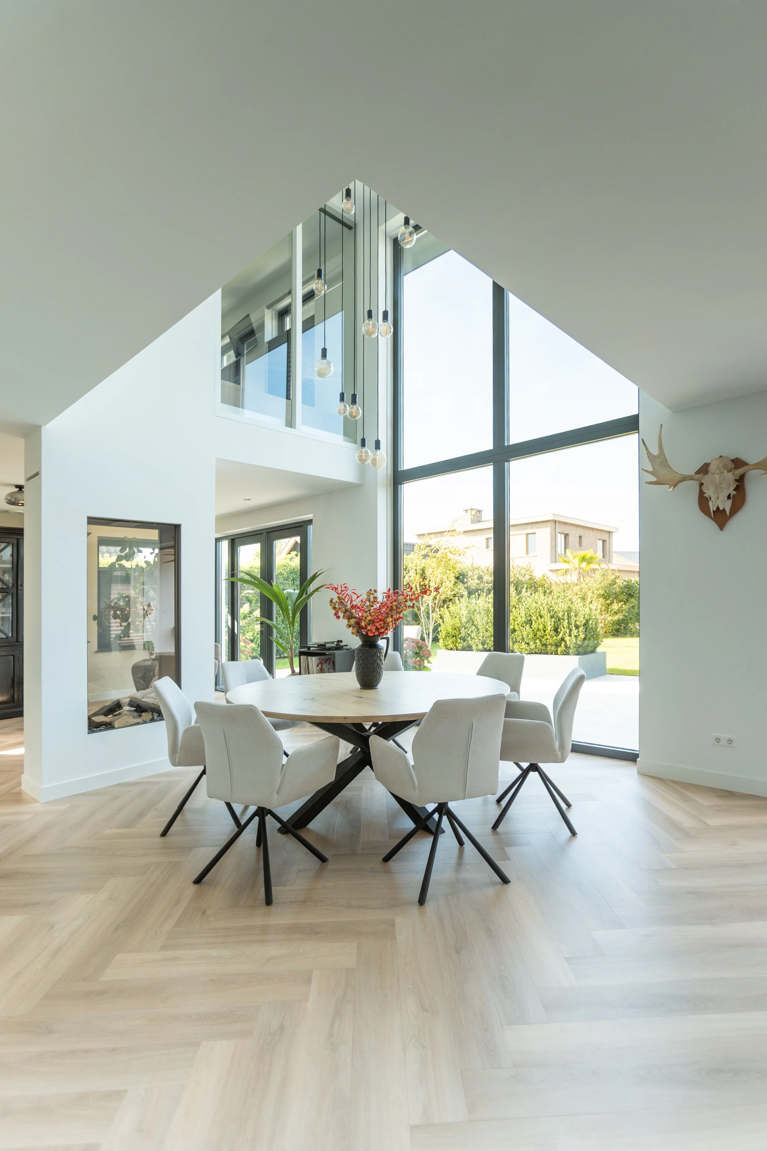 Eetkamer met wit ronde tafel, omgeven door acht witte stoelen op een houten vloer. Grote ramen en glazen deuren bieden uitzicht op een tuin. Decoratieve takken met bloemen en een gestileerde dierruif hangen aan de muur.