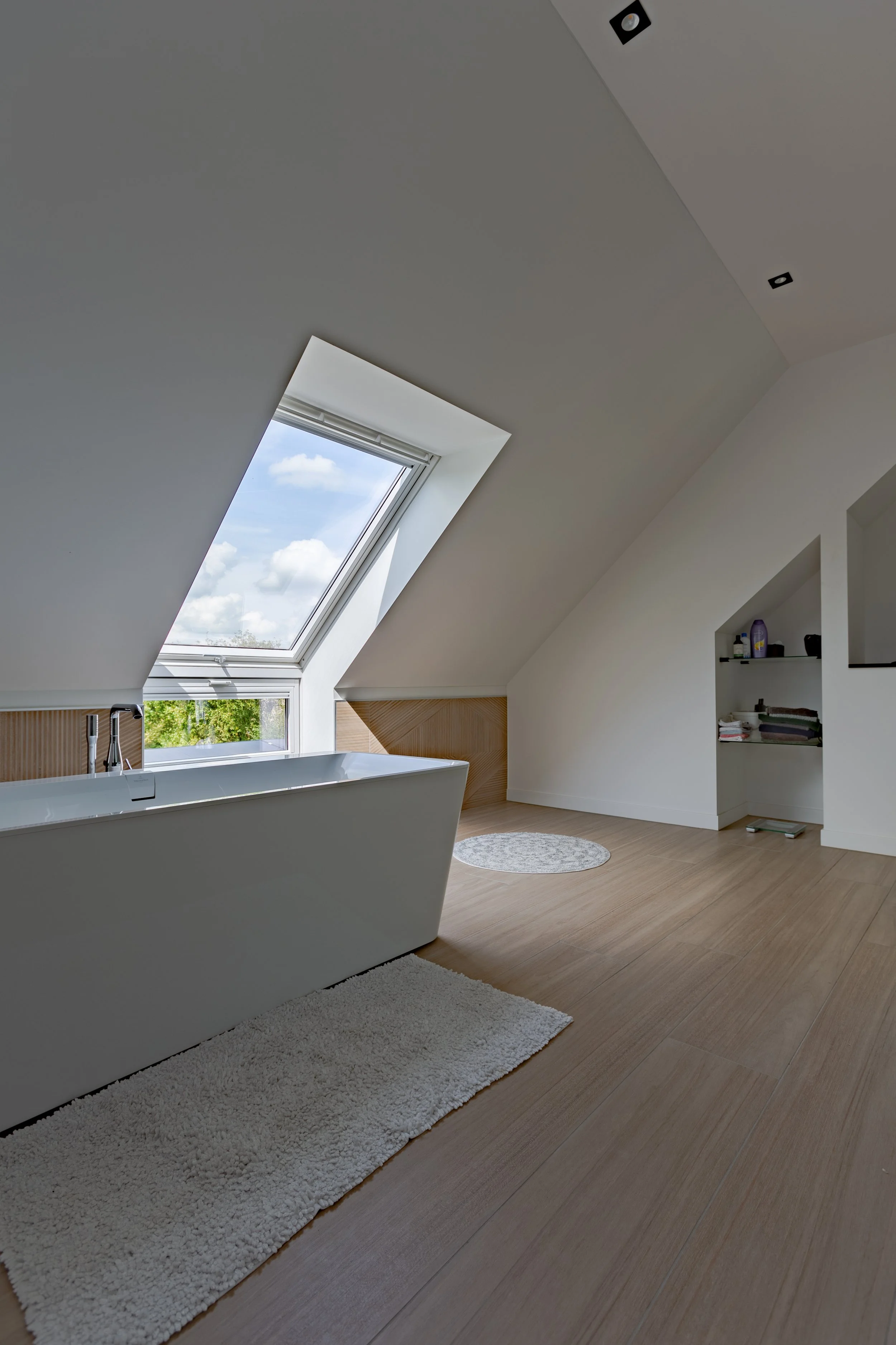 Een badkamer met een vrijstaand bad onder een grote dakraam dat uitzicht biedt op de blauwe lucht met witte wolken. De vloer is van licht hout, er ligt een witte spinnenweb-vormige badmat en er is een witte open kast met toiletartikelen.