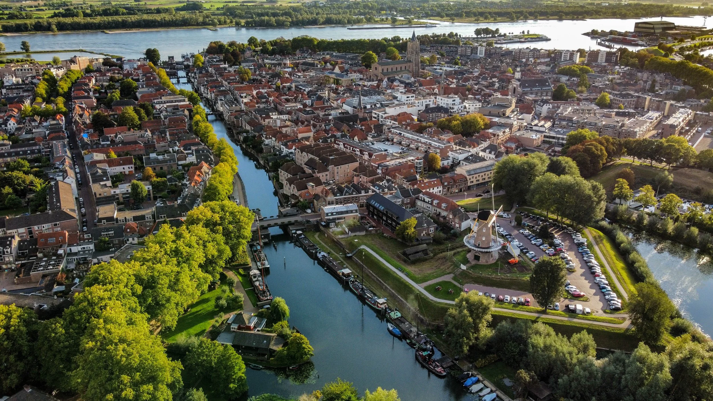 Een luchtfoto van een stad met een rivier die door het centrum stroomt, omgeven door groen en gebouwen, met een kerktoren en een windmolen te zien.