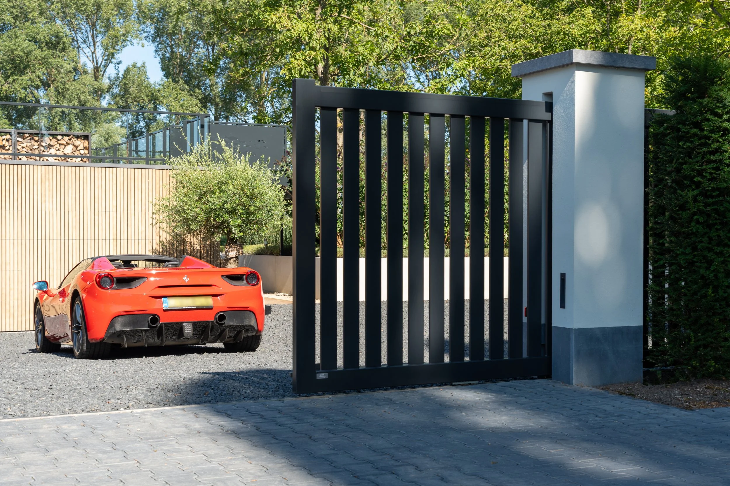 Een rode sportauto geparkeerd voor een zwart metalen poort en een witte betonnen pilaar, met groene bomen op de achtergrond.