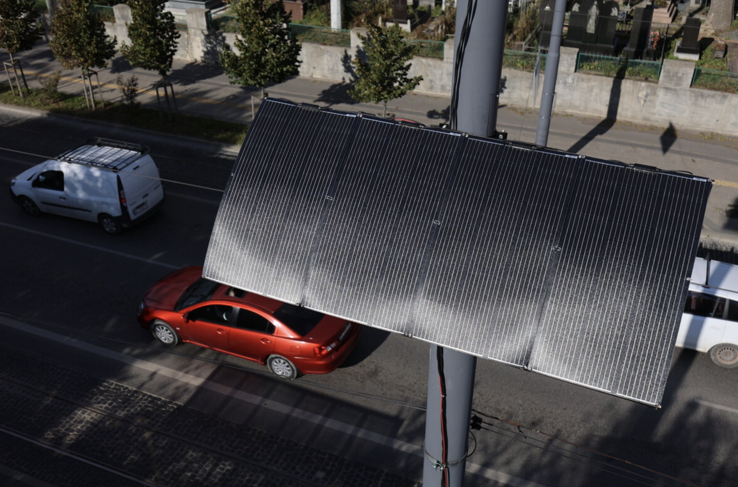 A red car parked on the street beneath a solar panel mounted on a pole. The street has trees and a sidewalk with a stone wall and fences in the background.