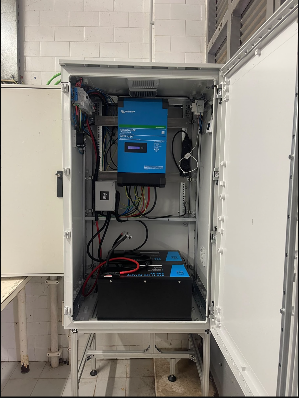 Electrical control cabinet with various wires, a large blue power inverter labeled 'EasySolar-II GX,' and a black battery pack at the bottom, all mounted inside an open metal enclosure in a utility room.