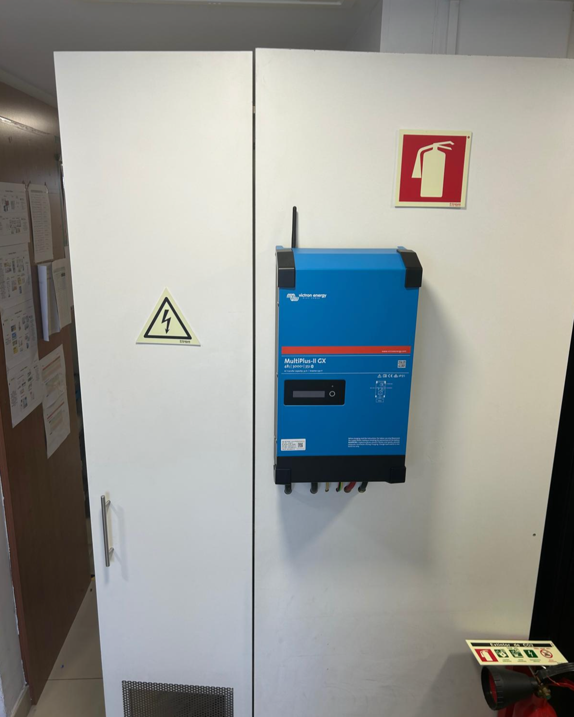 A white cabinet with warning and fire safety signs. A blue solar inverter is mounted on the cabinet, labeled 'MultiPlus-II GX' by Victron Energy. A fire extinguisher and safety instructions are partially visible at the bottom right.