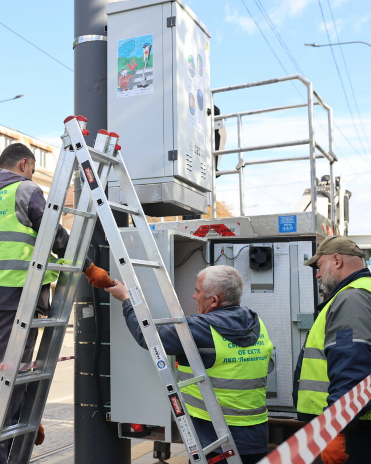 Workers installing or repairing electrical equipment on a utility pole using a lift platform and ladder in an outdoor urban setting.