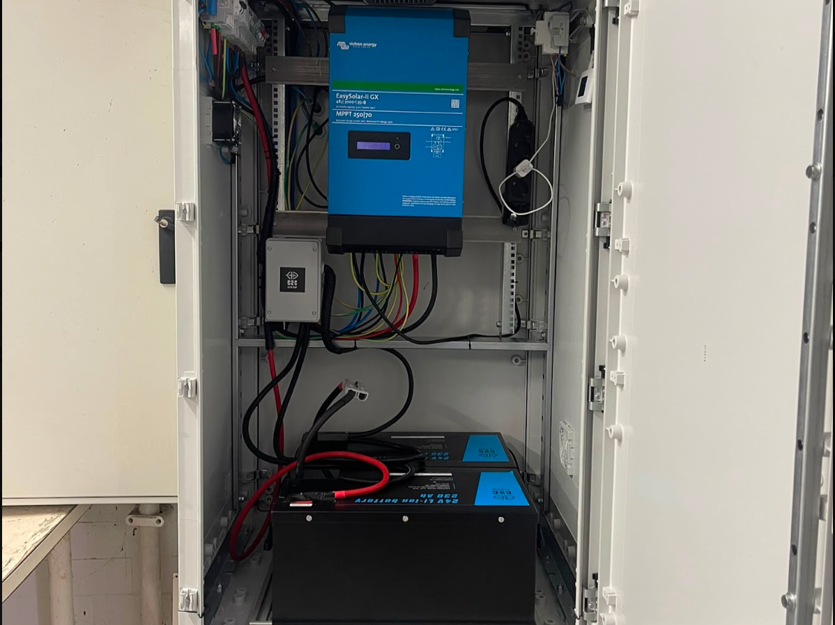 Interior of an electrical cabinet with various components, including a blue solar inverter and batteries connected with red and black cables.