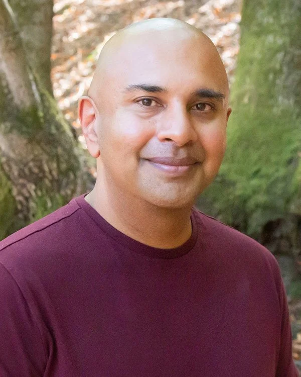 In this image Gullu is a bald man with a light complexion and a slight smile, wearing a maroon shirt, standing outdoors in front of trees with green moss and autumn leaves.