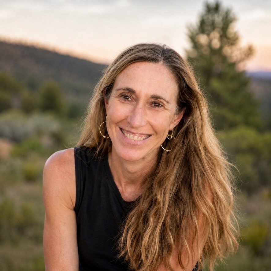 In this image Erin has long wavy hair and is smiling outdoors during sunset, wearing a black sleeveless top and hoop earrings.