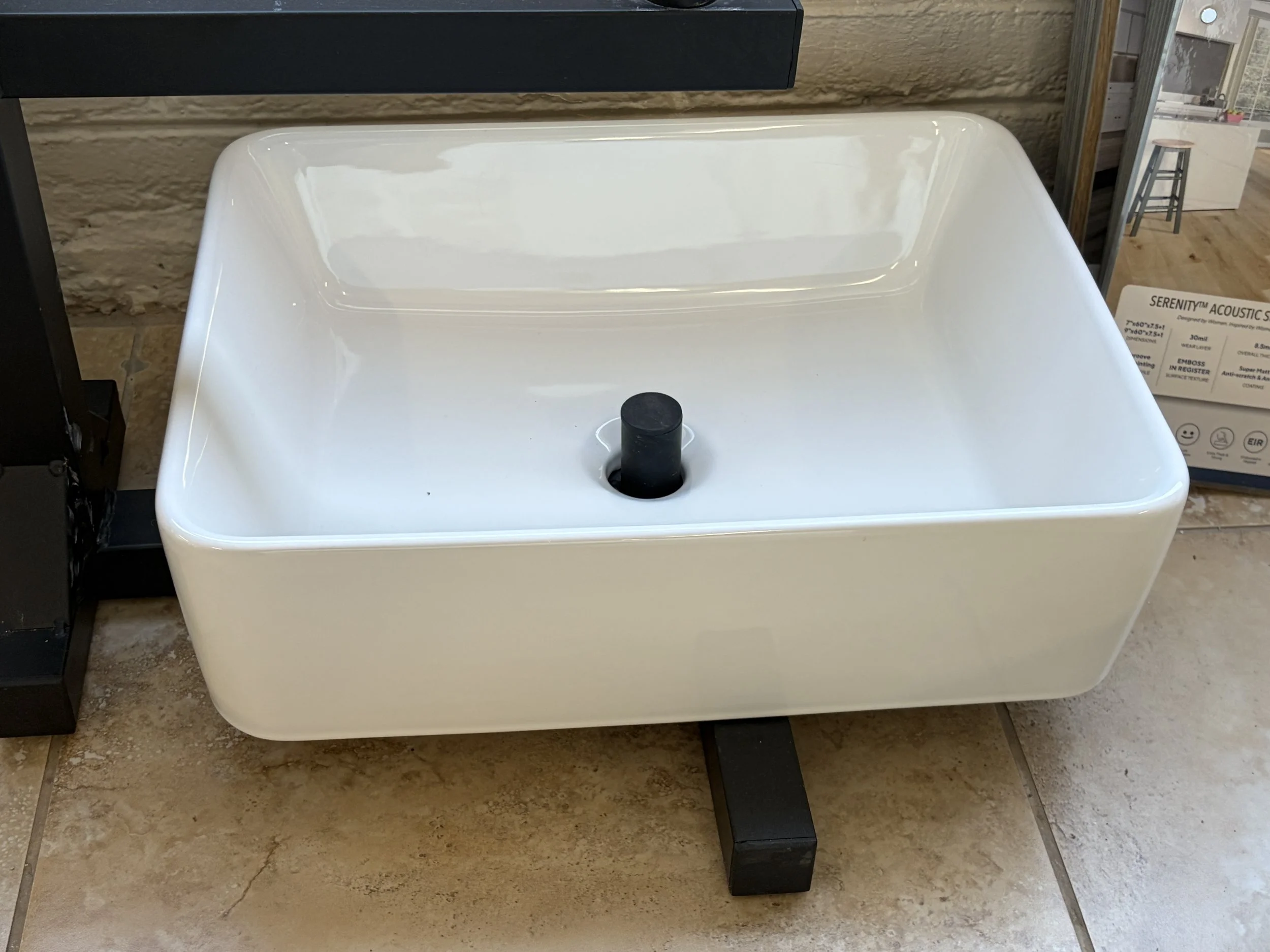 White rectangular bathroom sink with a black drain in the center, set on black legs, with a black shelf above it and a cardboard price tag to the right. The background features a beige textured wall and a tiled floor.