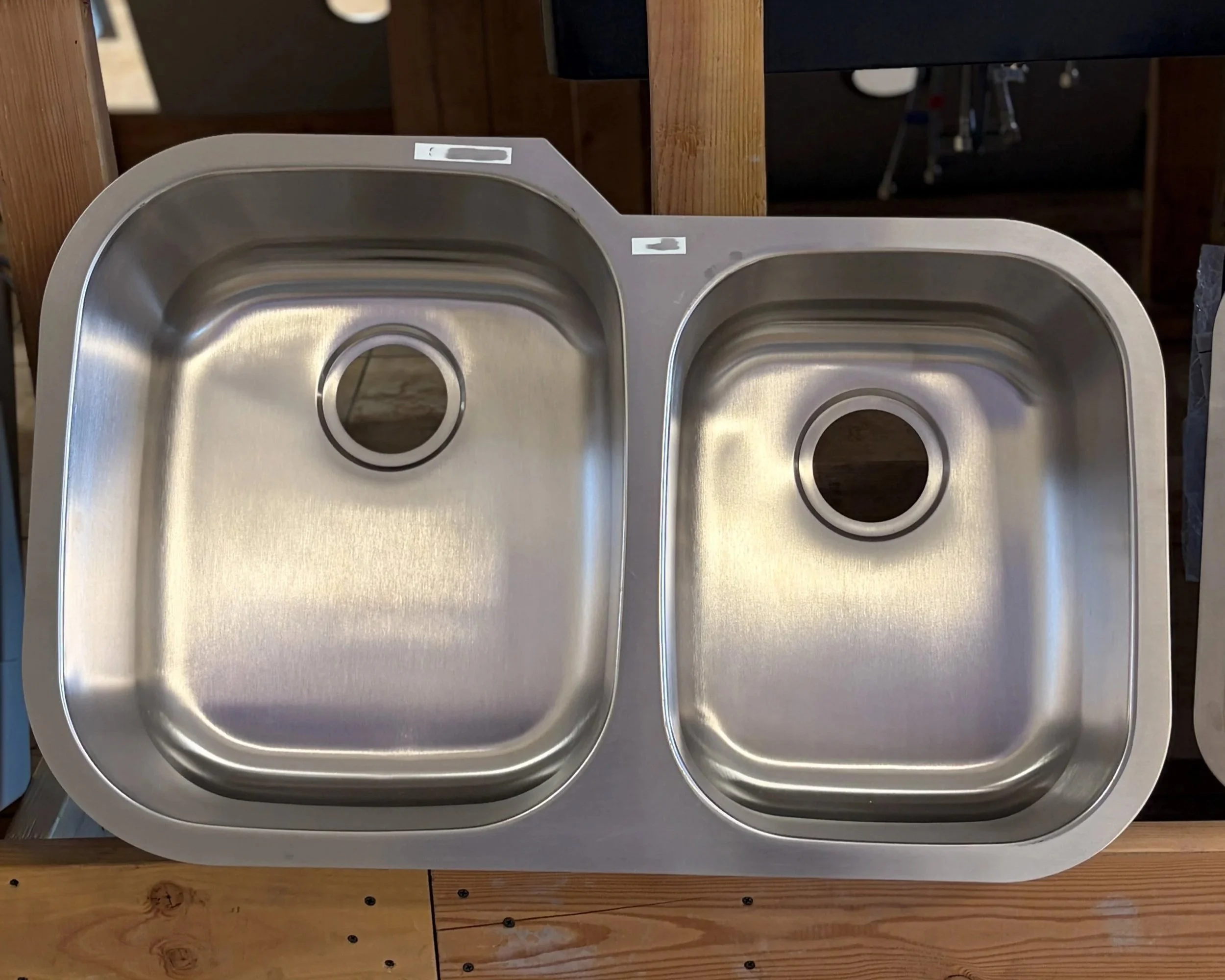 A stainless steel kitchen sink with two basins, placed on a wooden surface. The background features wooden beams and some kitchen or workshop tools.