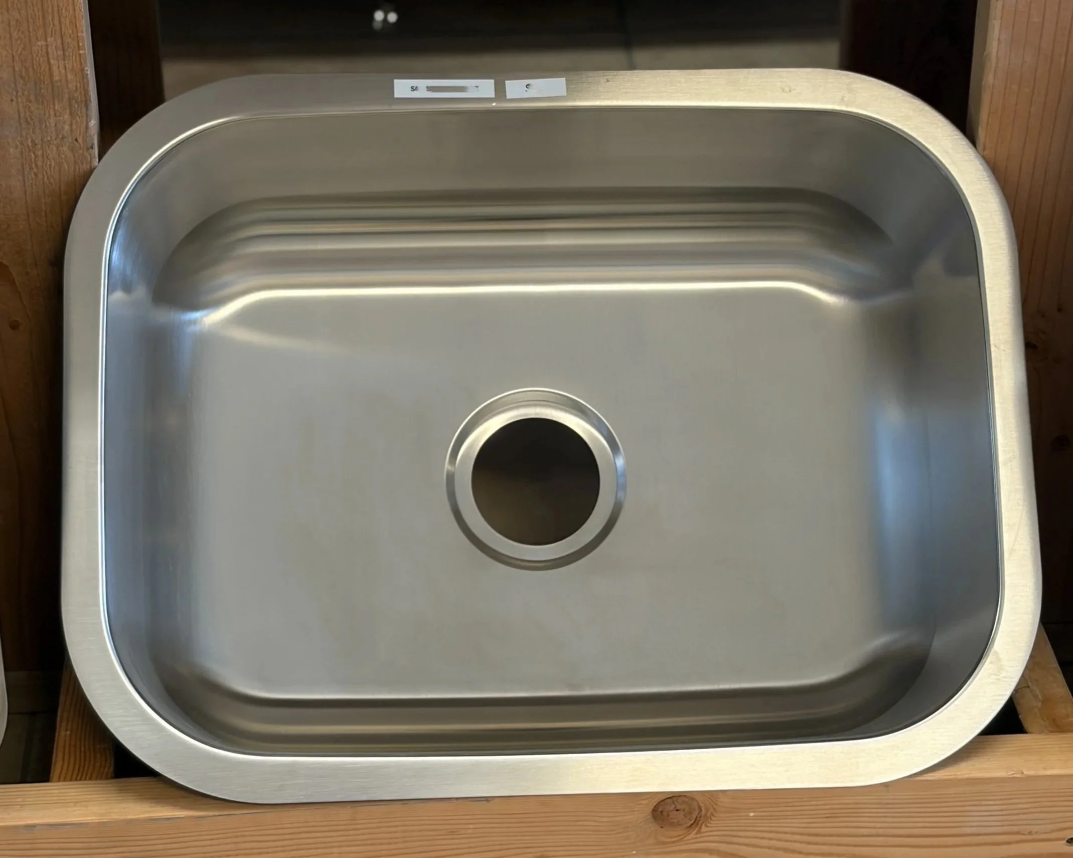 Close-up of a metallic, rectangular sink with rounded corners, mounted on a wooden countertop. The sink has a circular drain at the bottom center and is empty.