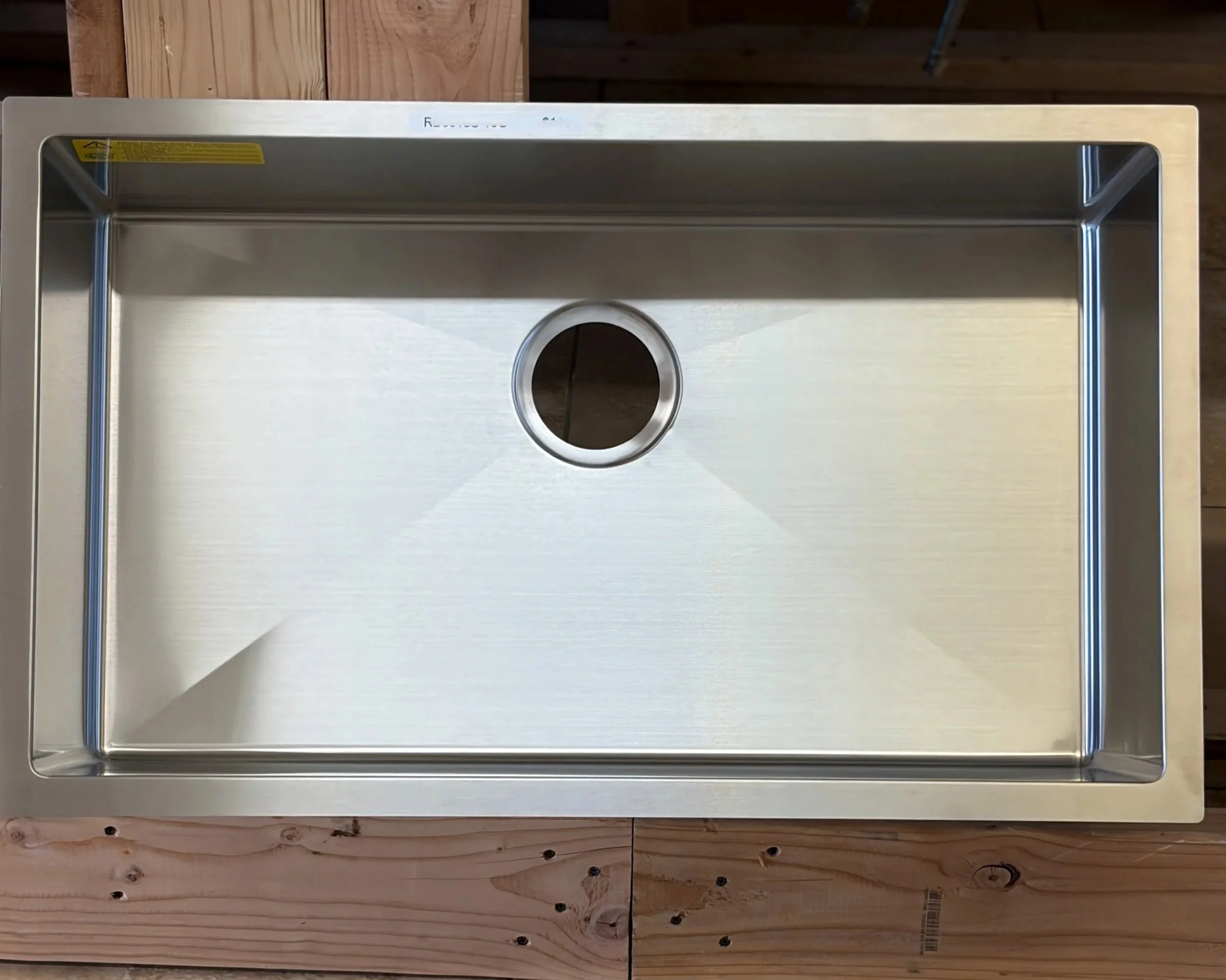 Stainless steel kitchen sink seen from above, installed on a wooden countertop. The sink has a circular drain opening in the center and rounded rectangular edges.