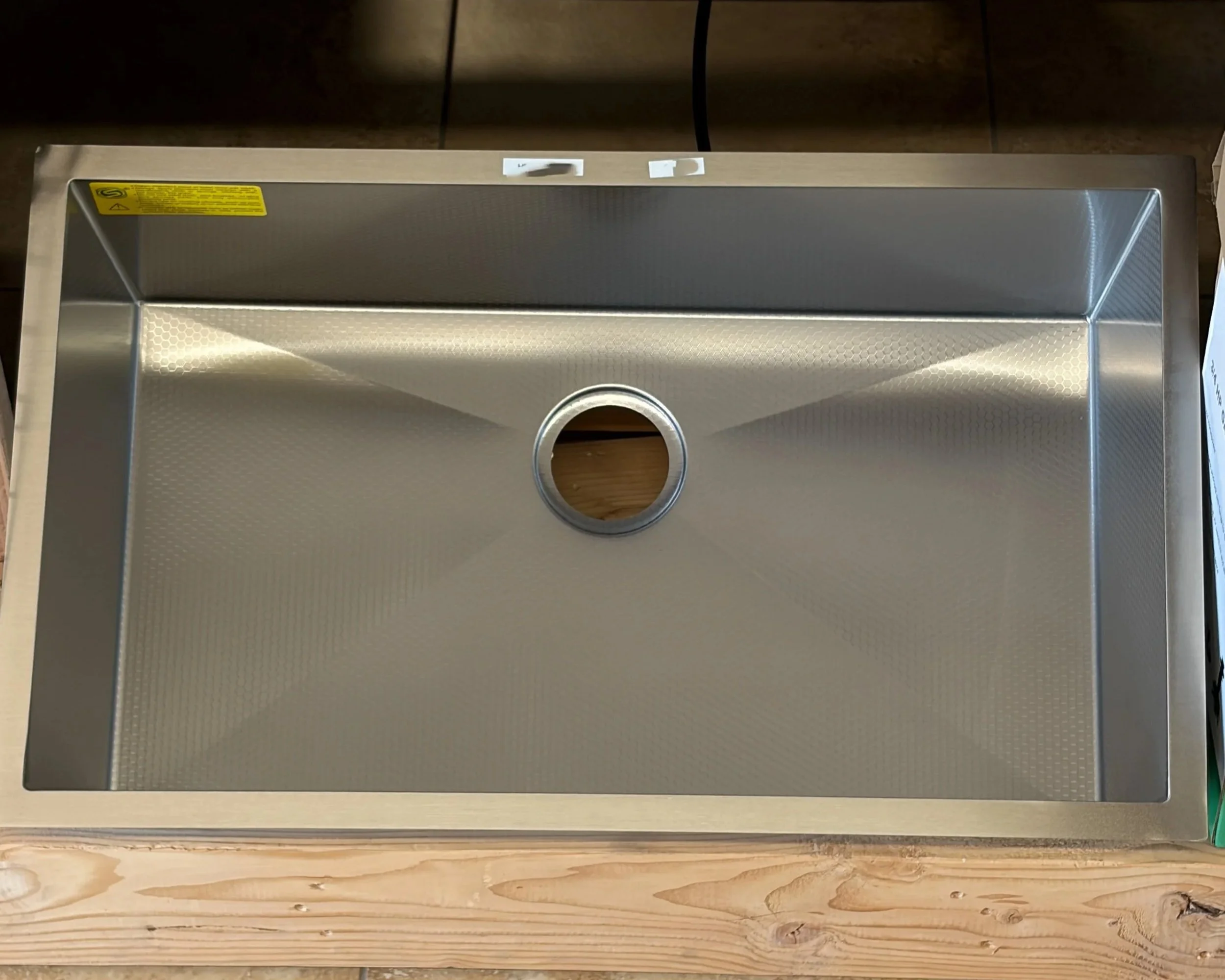 A stainless steel kitchen sink with a circular drain hole in the center, installed on a wooden countertop with a wood grain pattern.