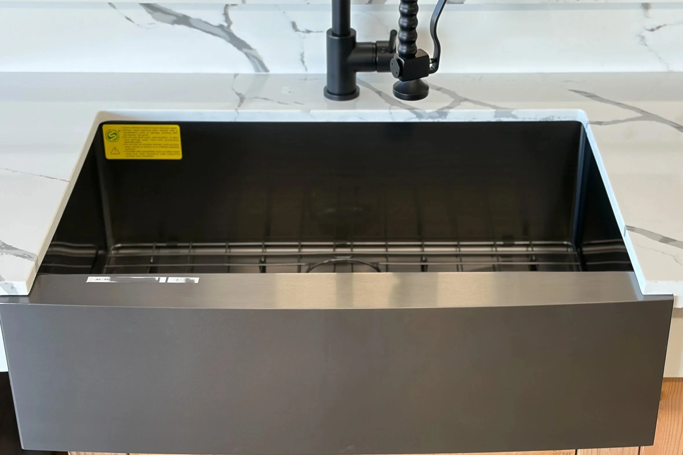 Empty kitchen sink with black faucet and marble countertop, with a yellow warning sticker on the metal edge of the sink.