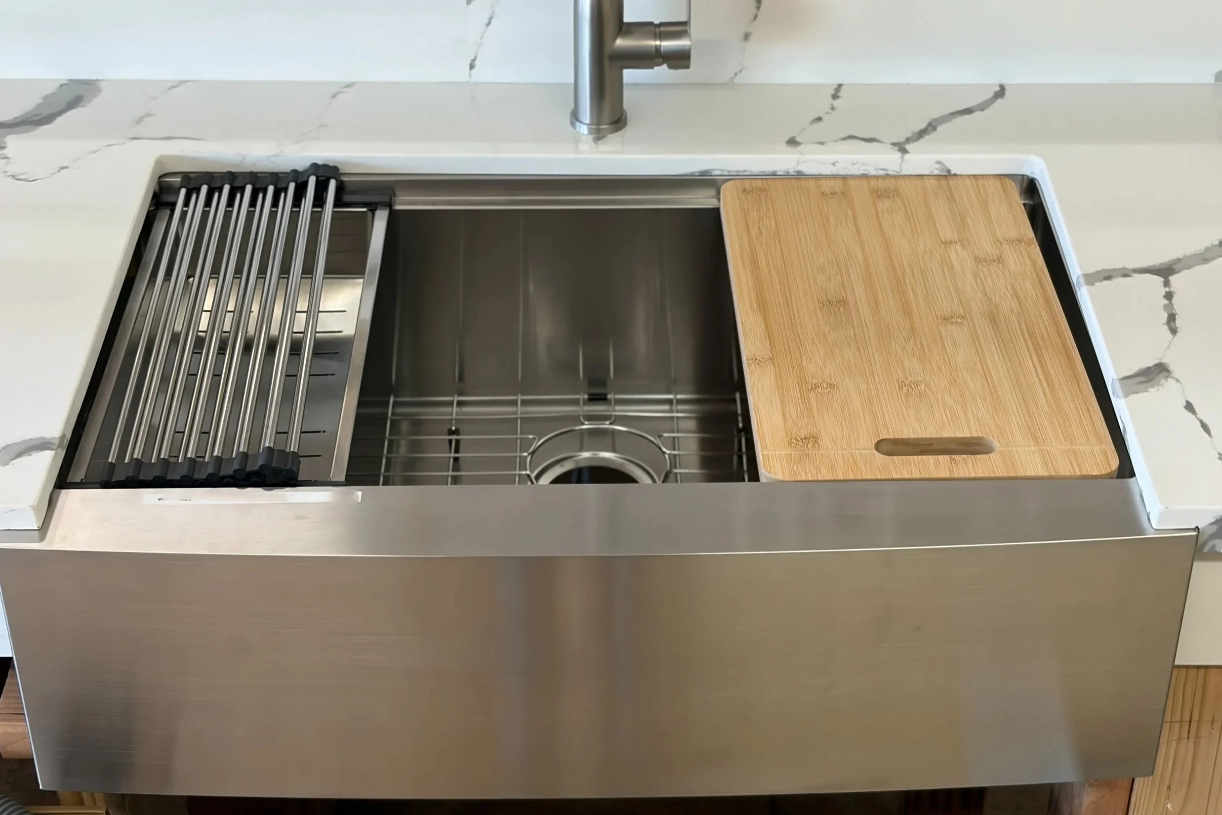 Top view of a kitchen sink with a stainless steel wire rack on the left side, a wooden cutting board on the right side, and a silver faucet above. The sink is set in a white marble countertop with gray veining.