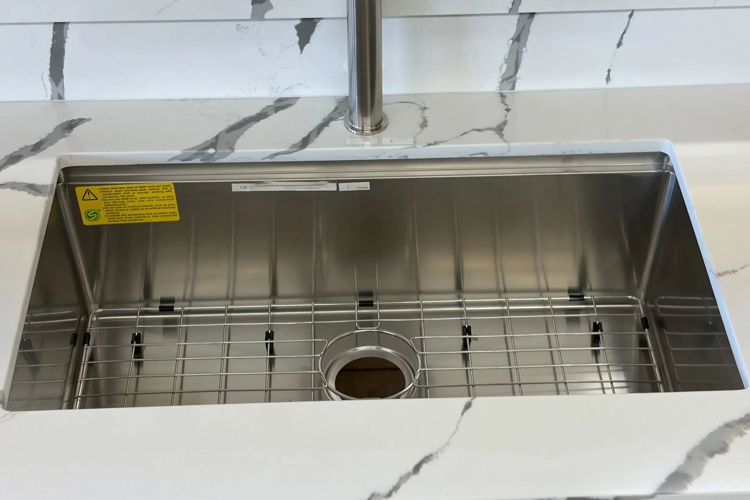 A stainless steel sink installed in a white marble countertop with gray veining, featuring a pop-up drain mechanism visible through the open drain opening.