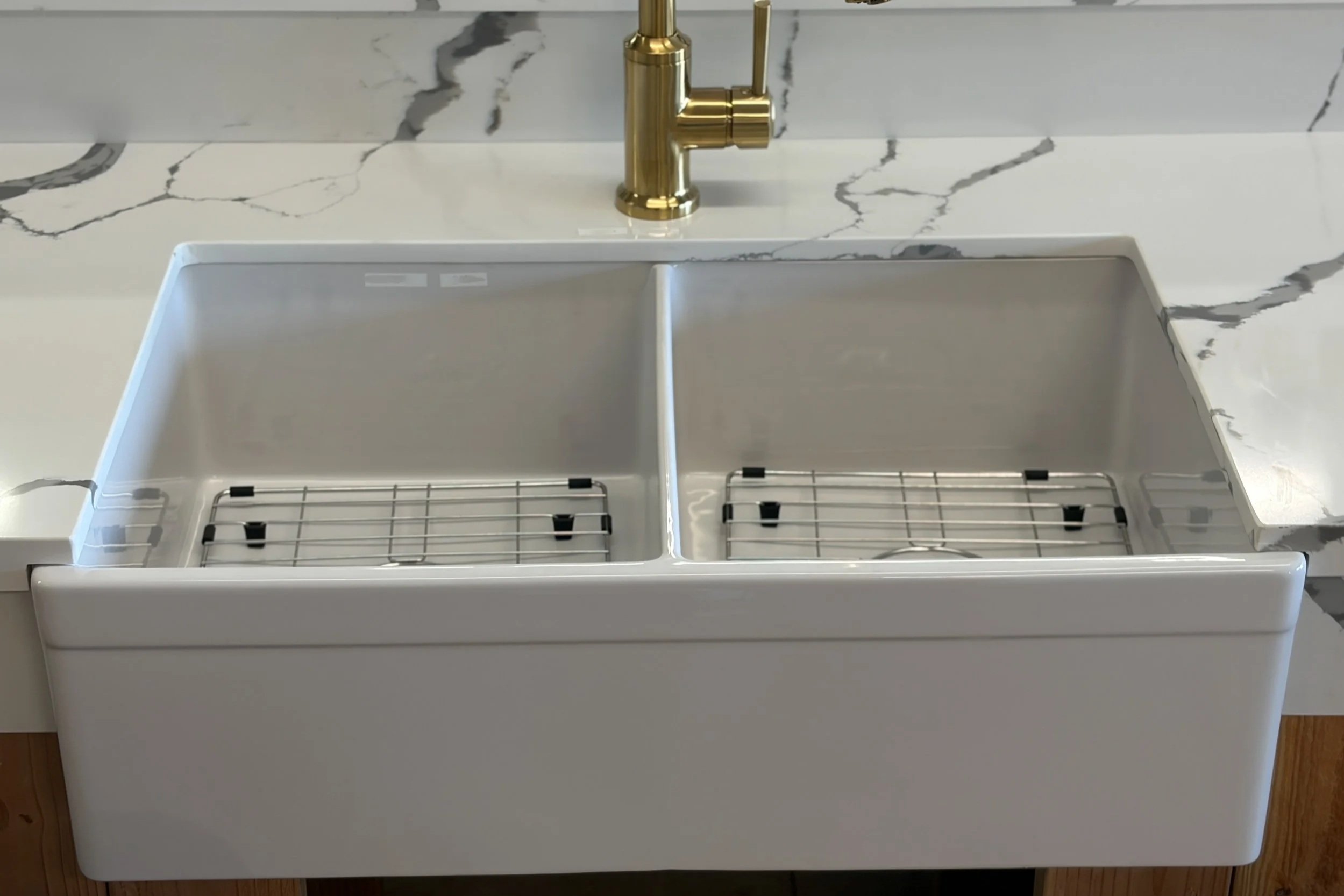 A white kitchen sink with a marble countertop and backsplash, featuring a gold faucet. The sink has a removable stainless steel dish drying rack inside.
