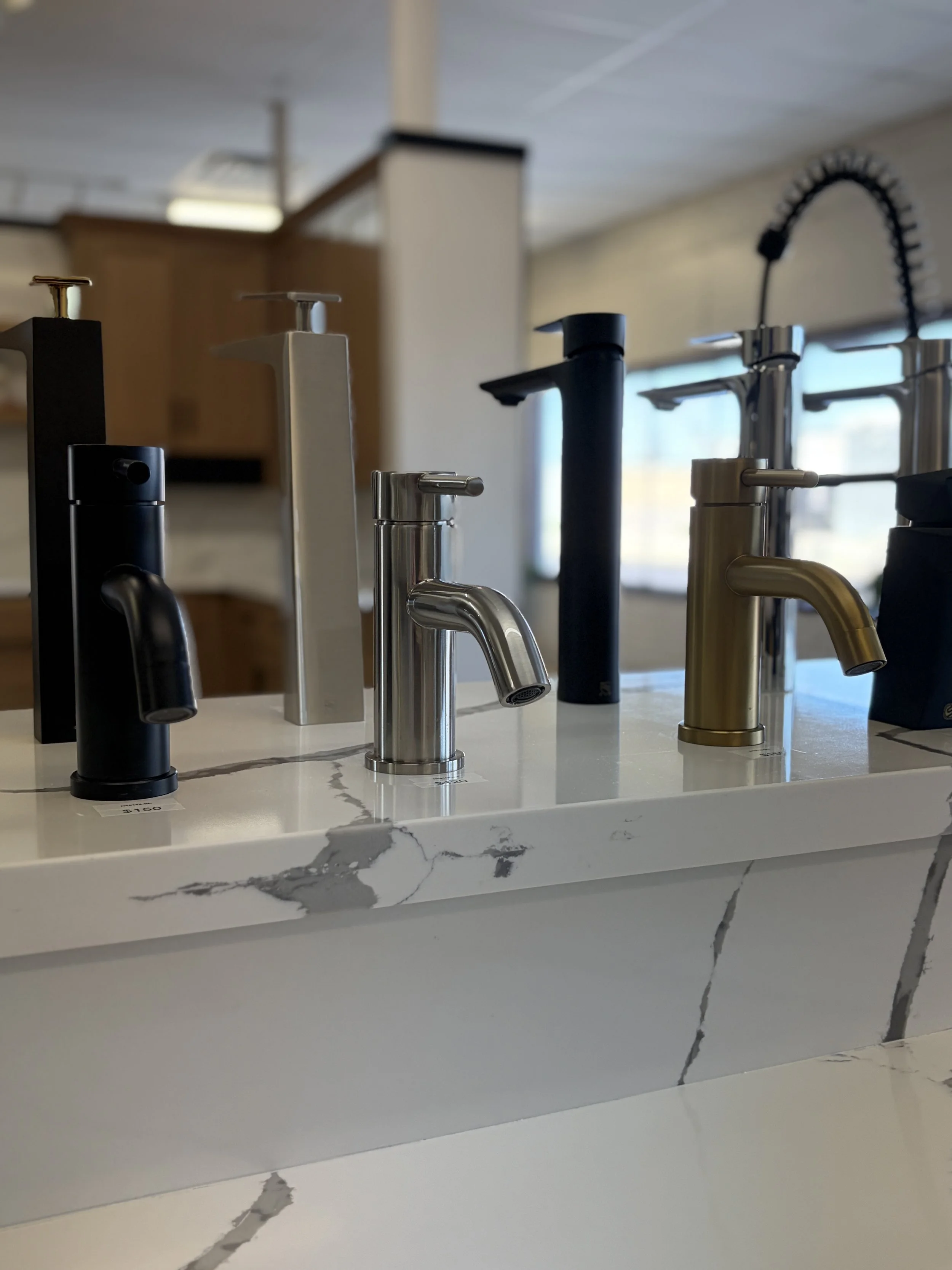 Display of various kitchen faucets in different finishes on a marble countertop.
