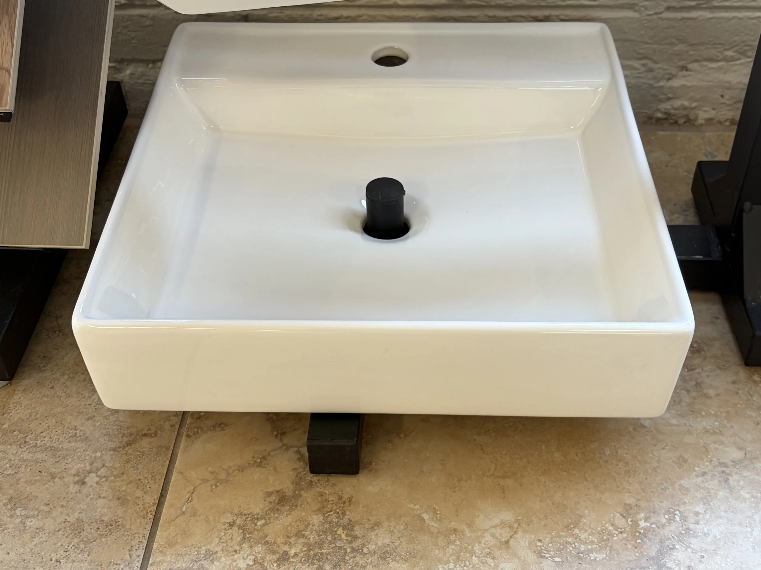 A white sink with a black drain stopper, placed on a black stand on a tiled floor.