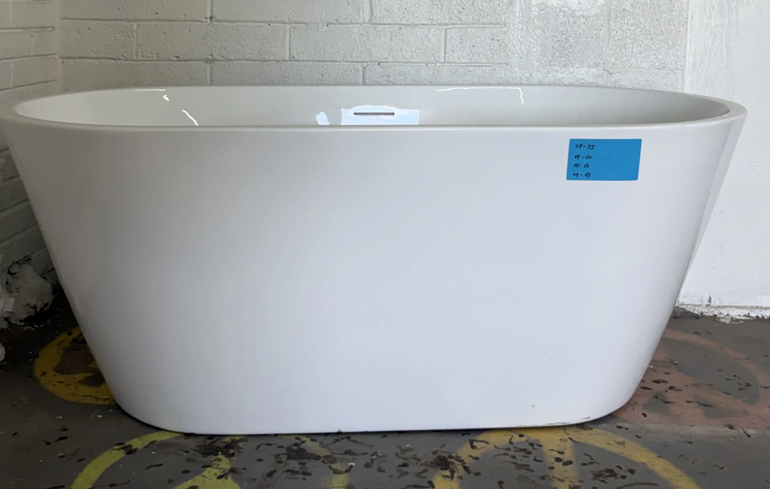 A white bathtub with a blue security tag attached to its side, situated in an unfinished room with concrete walls and a black and yellow safety line on the floor.